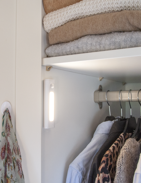 Armadio interno bianco con luce bianca illuminata. Scaffali con maglioni piegati, vestiti appesi sotto.