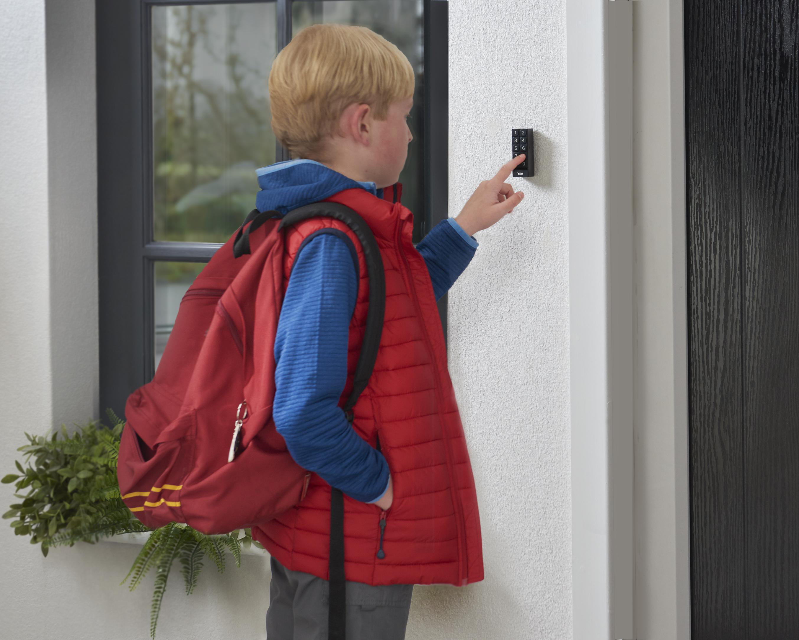 Blonder Junge in roter Weste mit Rucksack, der ein Tastenfeld an einer weißen Wand neben einer Tür benutzt.