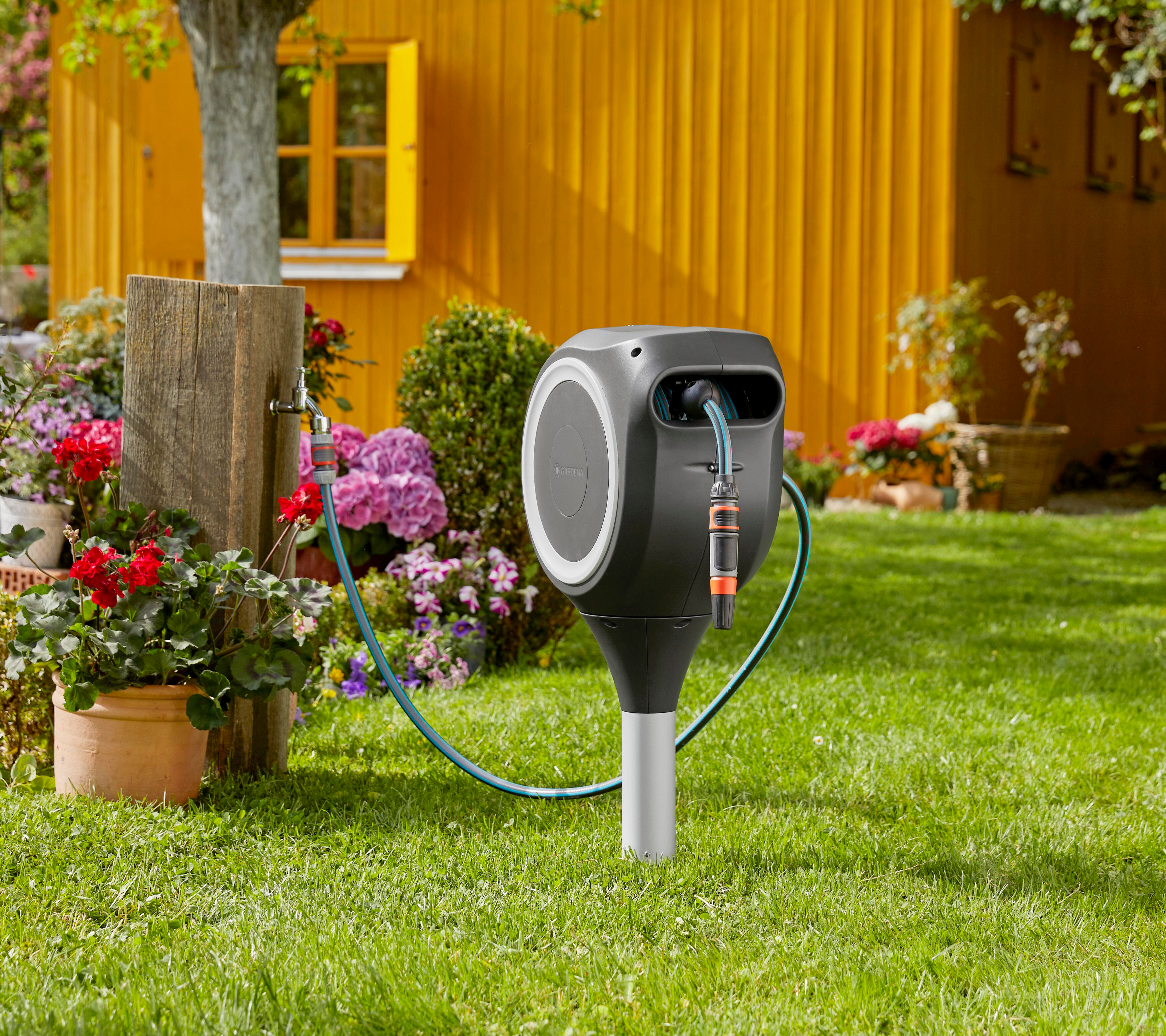 Eine Gartenszene mit einer Schlauchtrommel, Blumen und einem gelben Gebäude im Hintergrund.