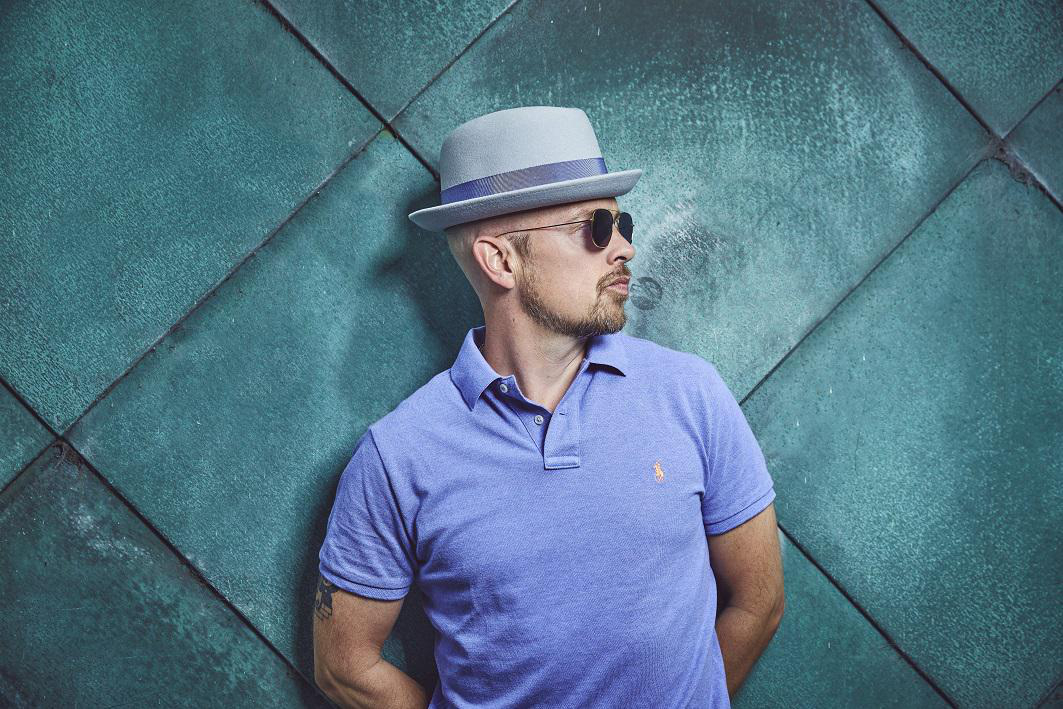 Mann in grauem Fedora, Sonnenbrille und blauem Poloshirt, lehnt an tealfarbener Wand.