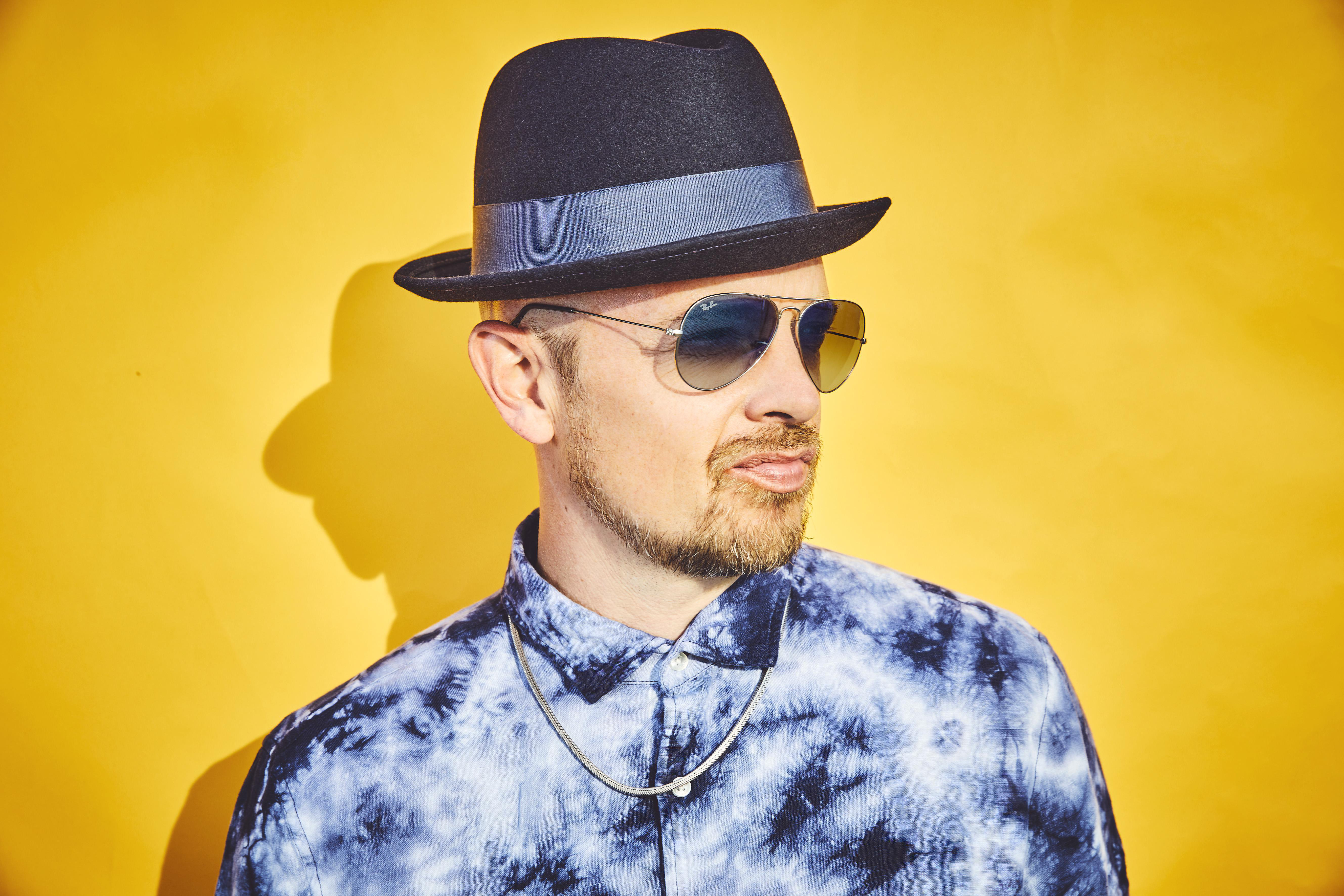 Ein Mann mit Hut und Sonnenbrille posiert vor gelbem Hintergrund.