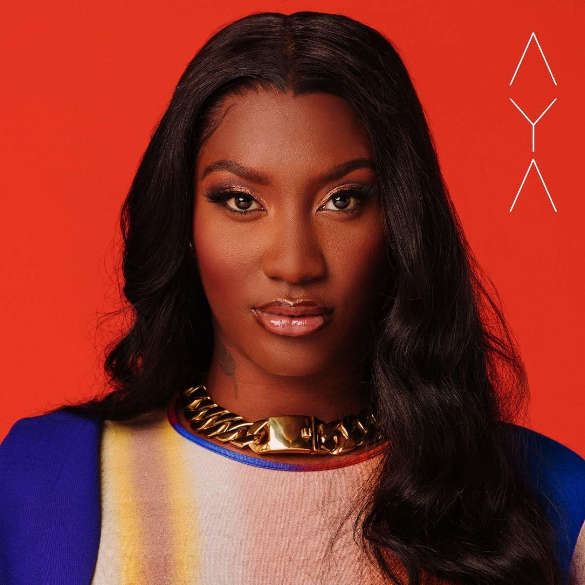Aya, avec de longs cheveux noirs, portant un collier doré, sur fond rouge. Logo triangle blanc au-dessus.
