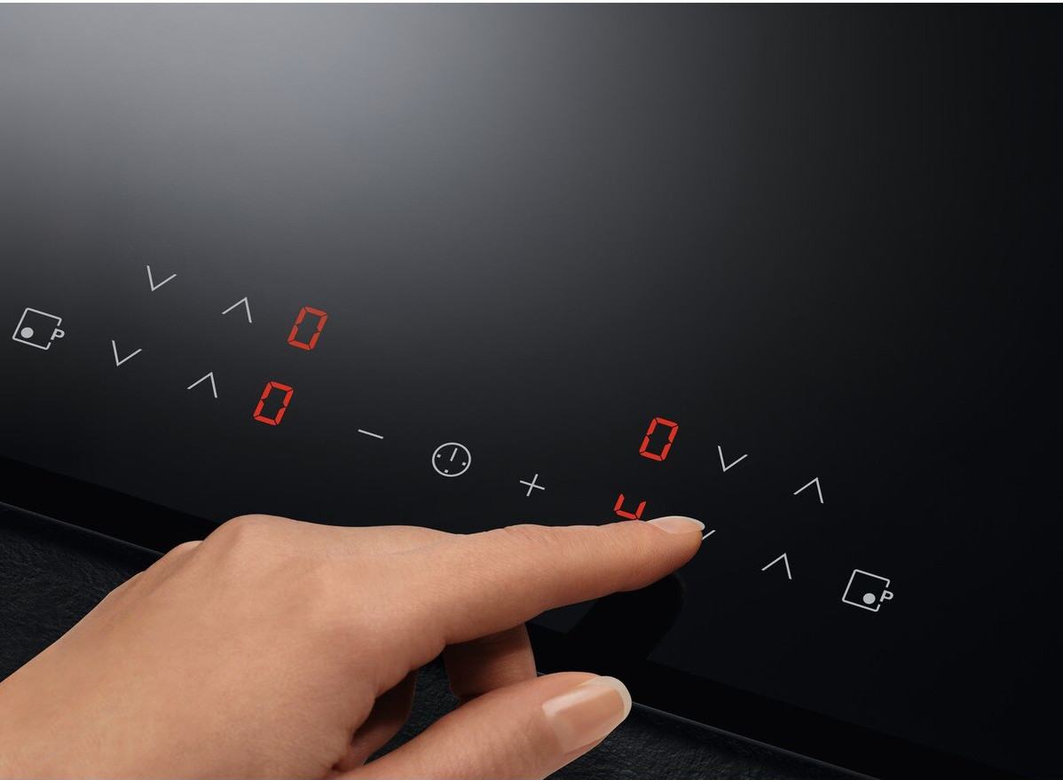 Hand touching a black cooktop. Red digits are displayed. Black background.