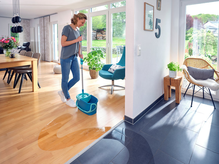 Een vrouw die een natte houten vloer dweilt. Een deel van de vloer is al schoon en droog.