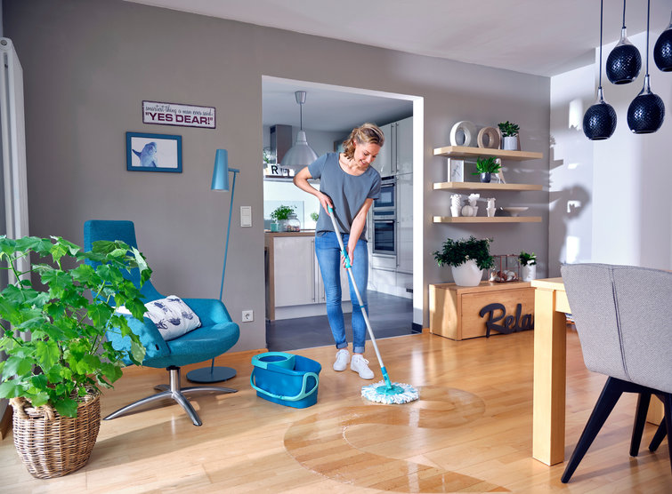 Vrouw die een houten vloer dweilt in een woonkamer. Emmer en dweil in gebruik. Grijze muren en blauwe accenten.