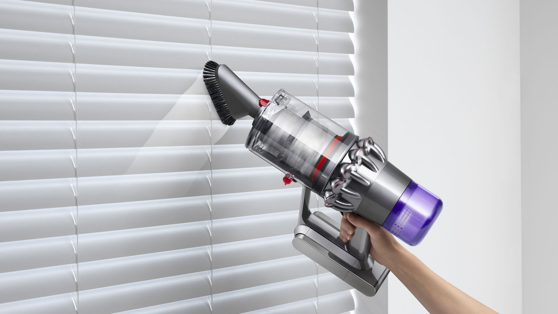 Gümüş ve mor bir Dyson elektrikli süpürge ile beyaz panjurları süpüren bir kişi.