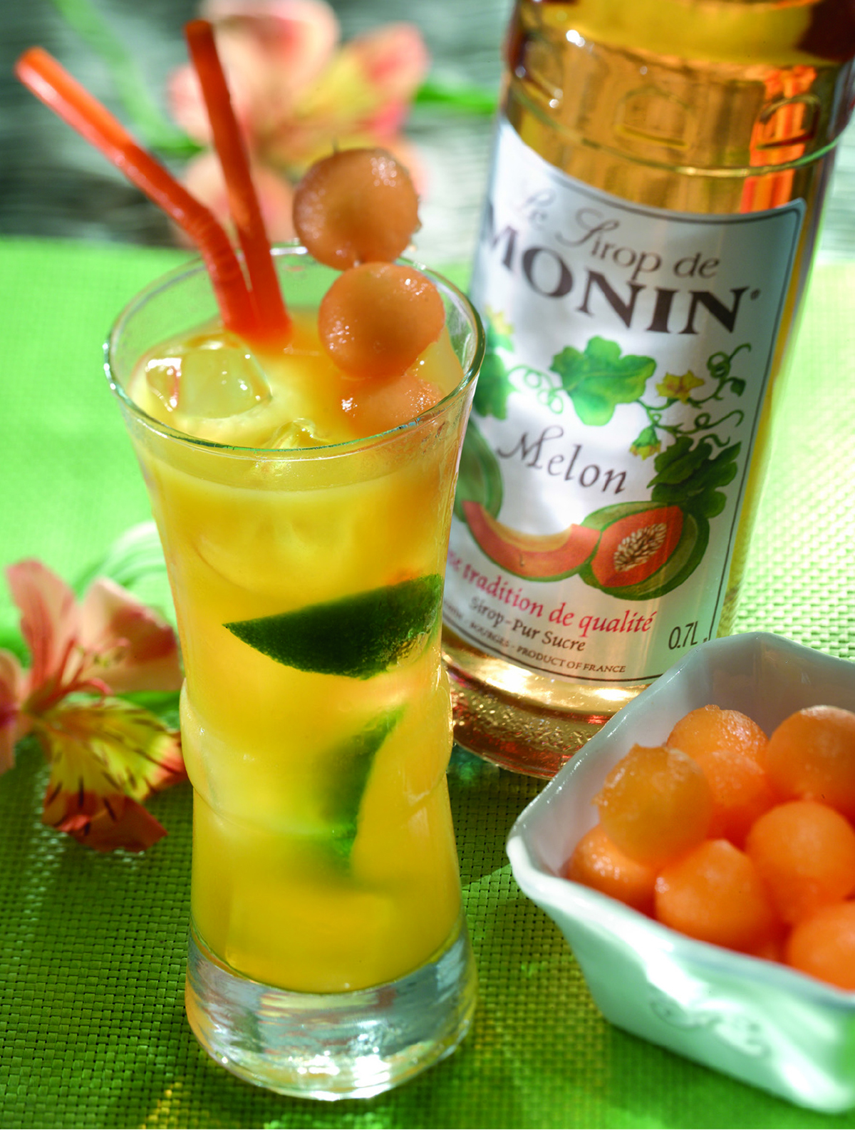 Hohes Glas Melonen-Cocktail mit Eis, Melonenbällchen und eine Flasche Monin Melonen-Sirup im Hintergrund.
