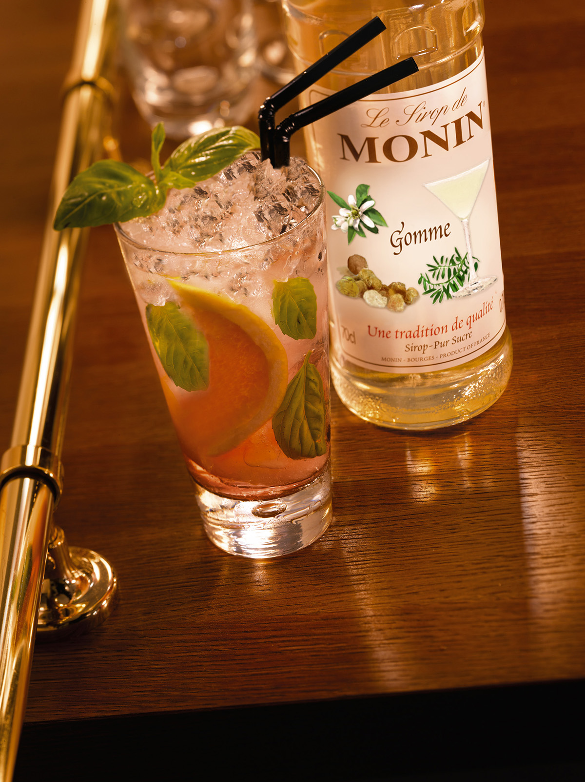 Ein Cocktail im Glas mit Eis, einer Zitronenscheibe und Minzblättern. Eine Monin Sirup Flasche ist im Hintergrund.