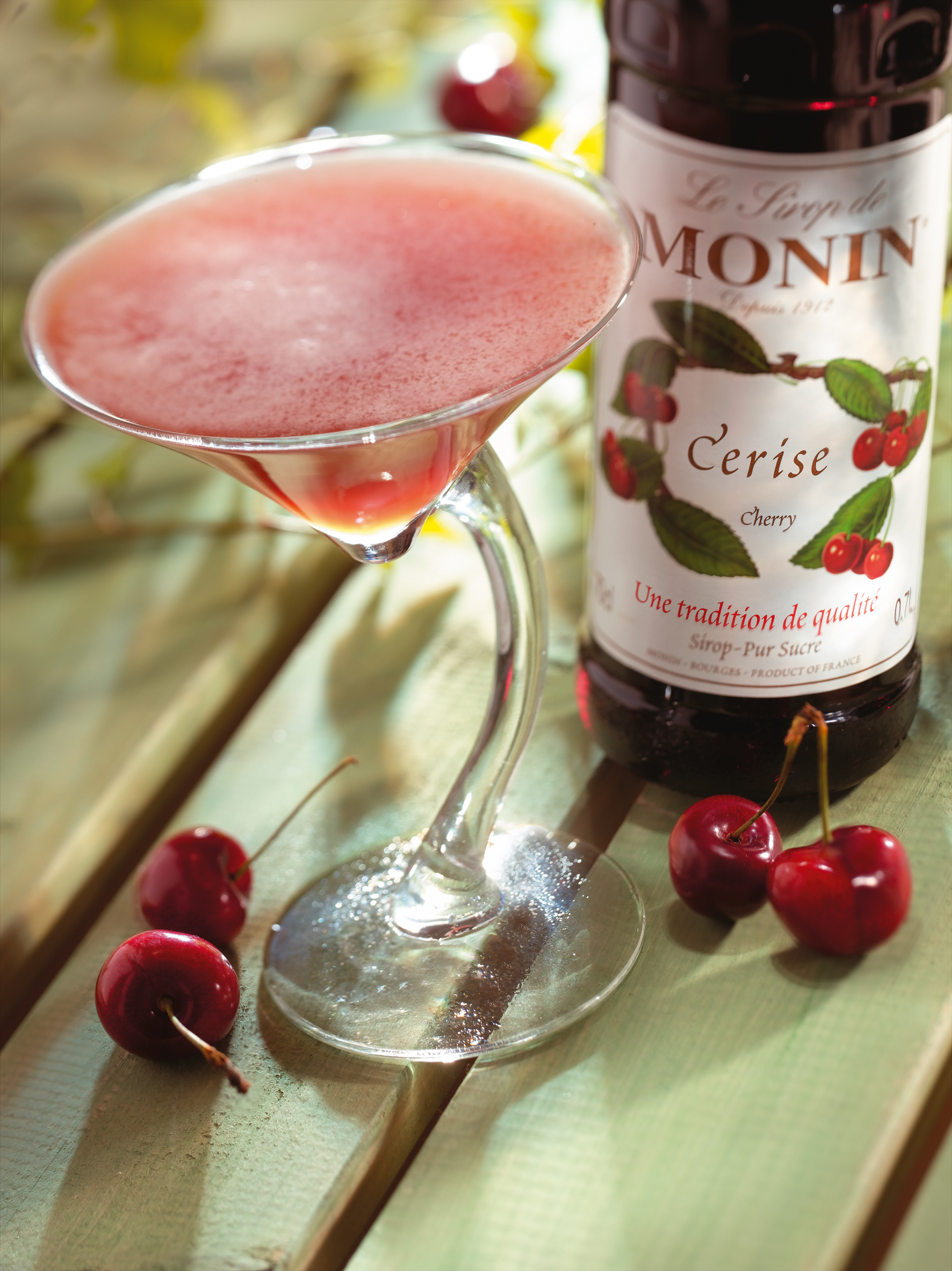 Kirschcocktail mit einer Monin Sirupflasche und Kirschen. Grüne Holzoberfläche.