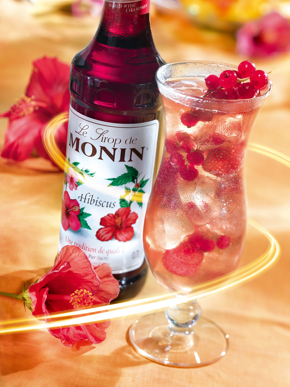 Ein Cocktail mit roten Beeren und Eiswürfeln, mit einer Flasche Hibiskus-Sirup und einer Hibiskusblüte.