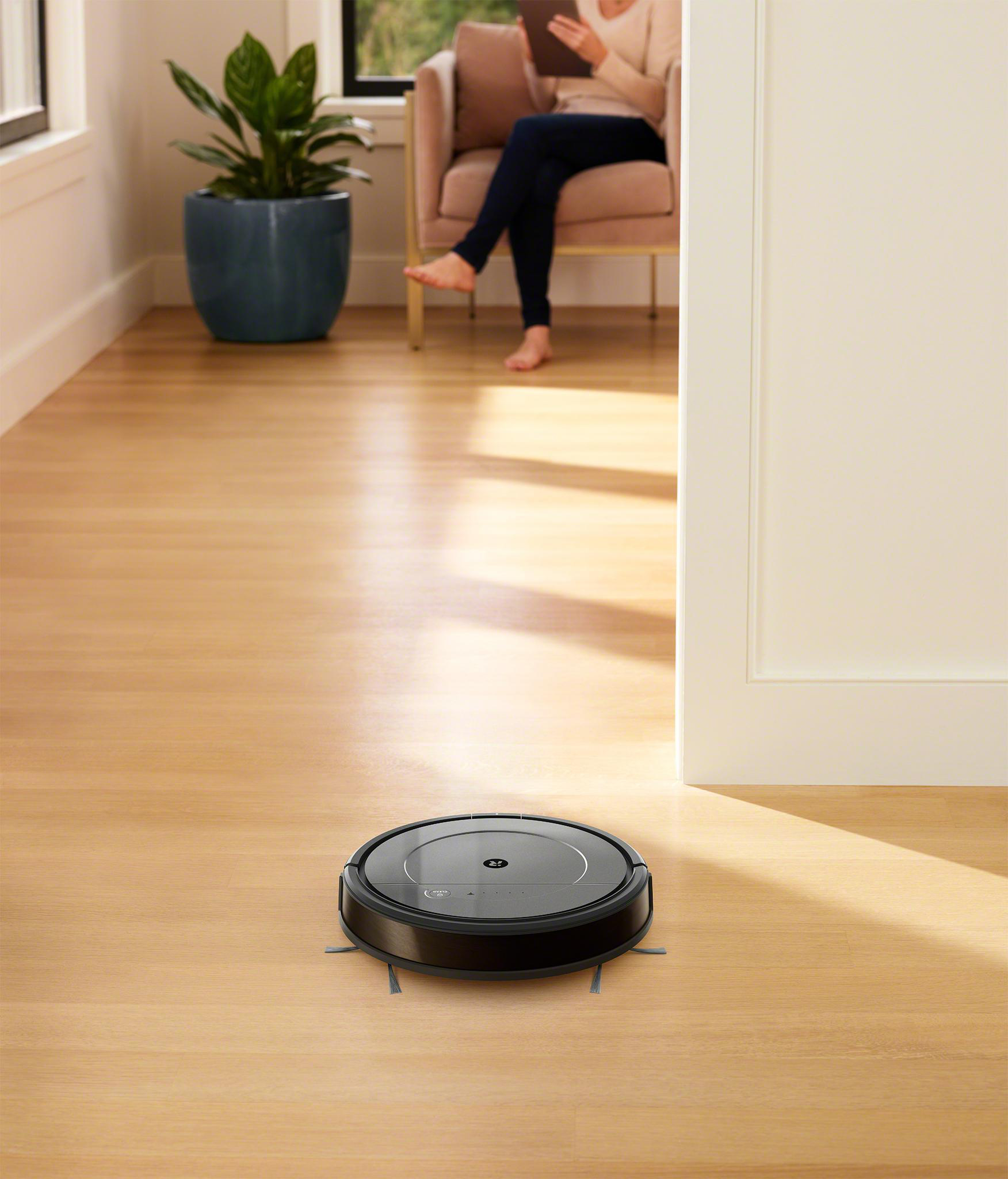 Roboter-Staubsauger auf Holzboden. Eine Frau sitzt im Hintergrund auf einem Sofa und liest.