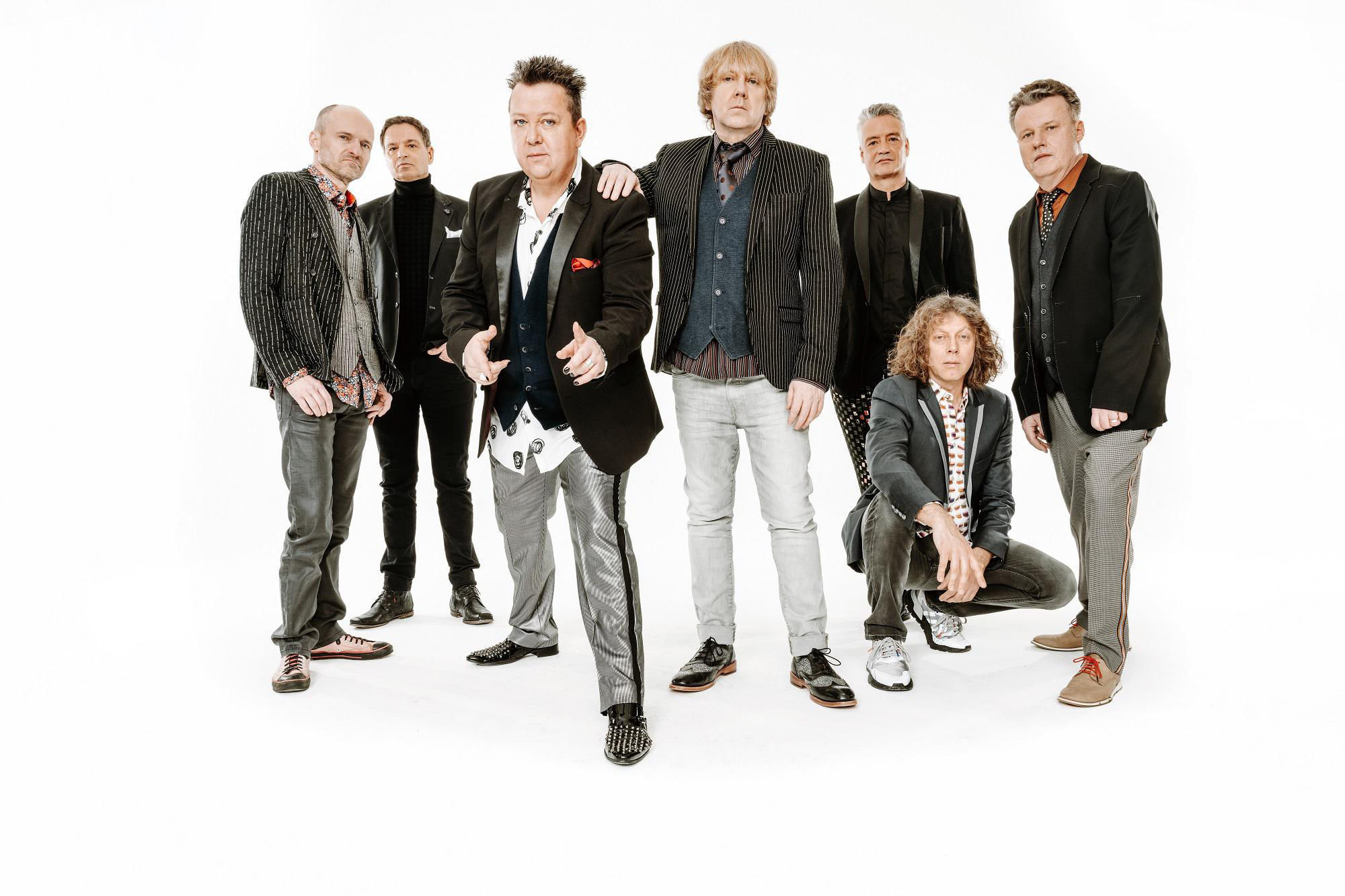 Siebenköpfige Band, die vor weißem Hintergrund posiert und verschiedene Outfits trägt.
