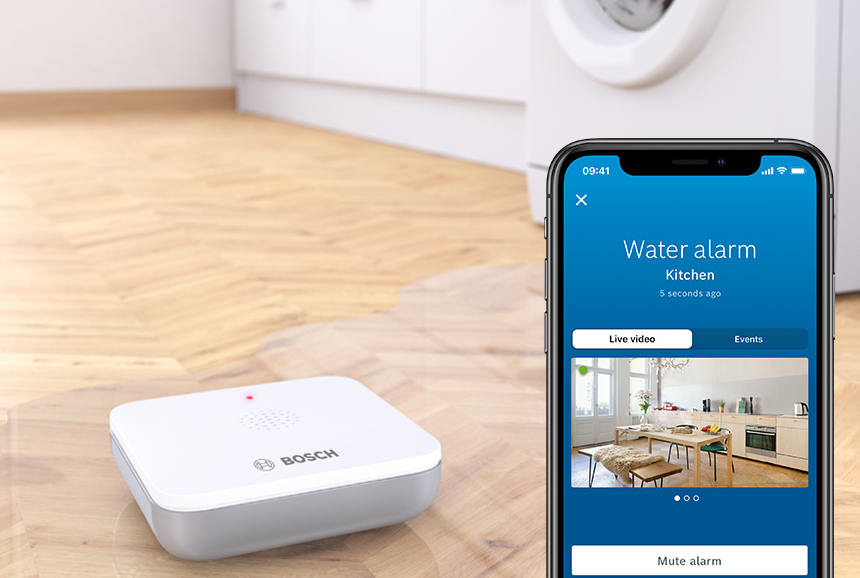 Ein Wasseraustrittssensor auf einem Boden mit einem Telefon, das einen Wasseralarm in einer Küche mit Live-Video zeigt.