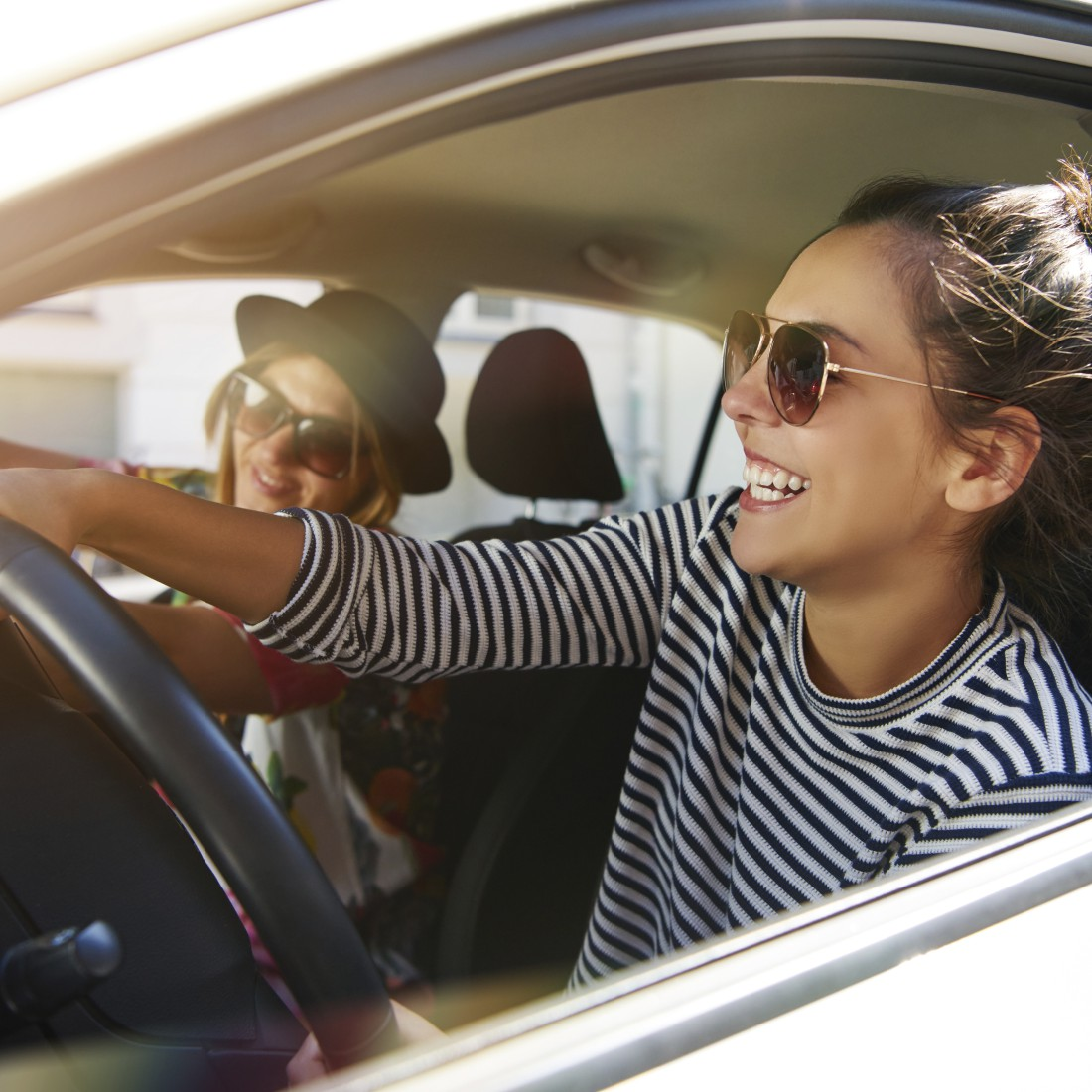 Zwei Frauen in einem Auto, eine fährt und lächelt, die andere trägt eine Sonnenbrille.
