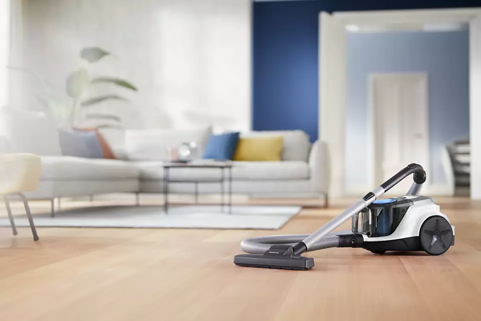 Ein Staubsauger auf Holzboden in einem Raum, mit Sofa und blauer Wand im Hintergrund.
