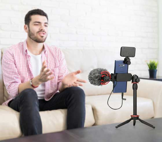 Un homme enregistre une vidéo avec un micro et un téléphone sur un trépied. Le fond est une pièce.