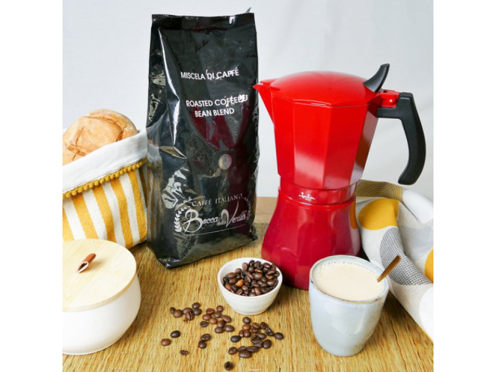 Varios artículos de café: bolsa, cafetera moka roja, taza, granos y pan sobre una mesa de madera.