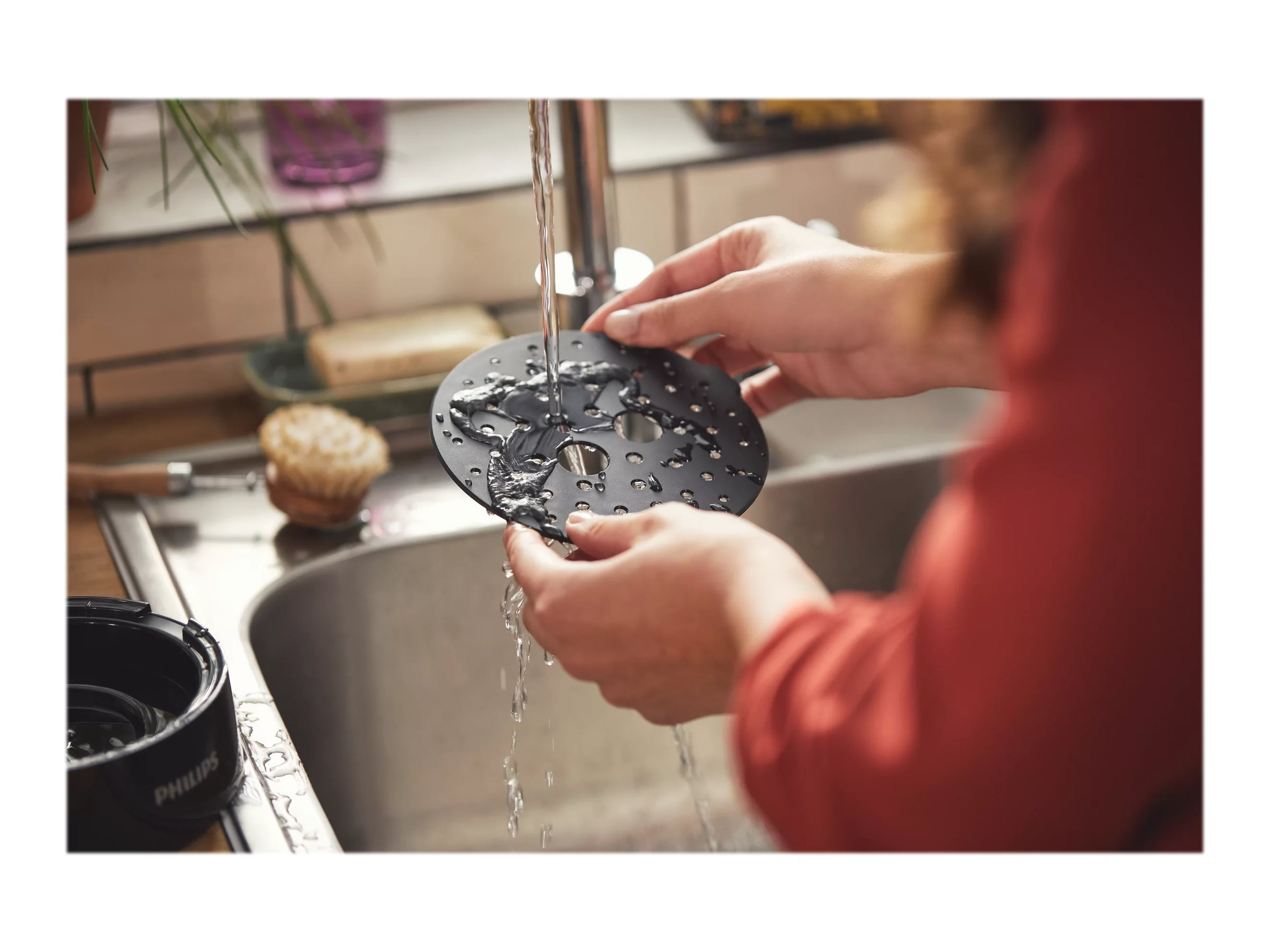 Hände spülen ein schwarzes Airfryer-Teil unter fließendem Wasser ab.