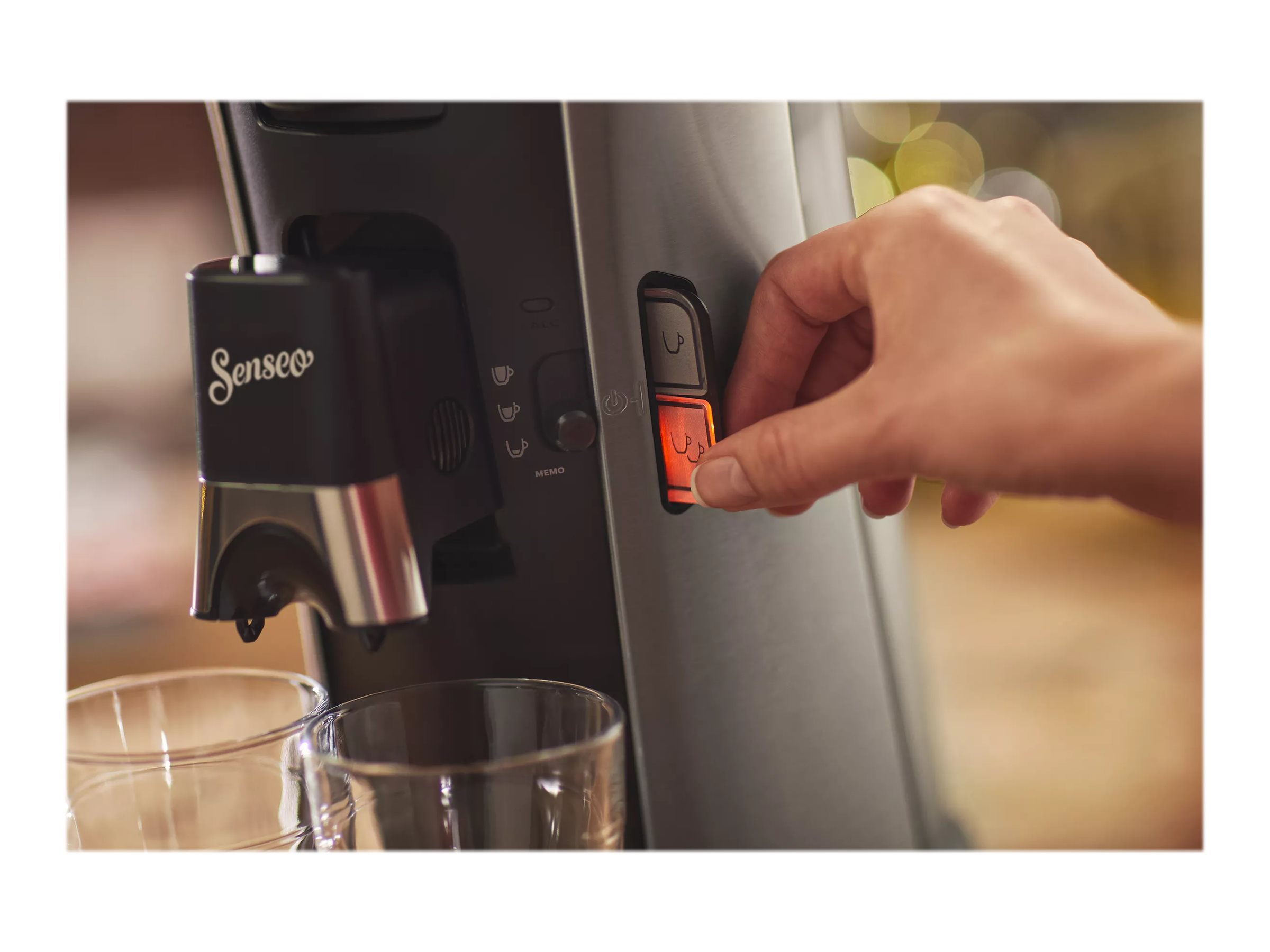Eine Hand drückt eine Taste an einer schwarzen Kaffeemaschine, Gläser sichtbar.