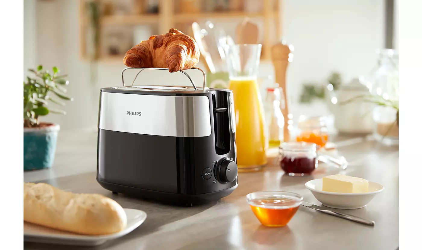 Egy fekete-ezüst Philips kenyérpirító croissant-val a tetején és reggeli elemekkel körülvéve.