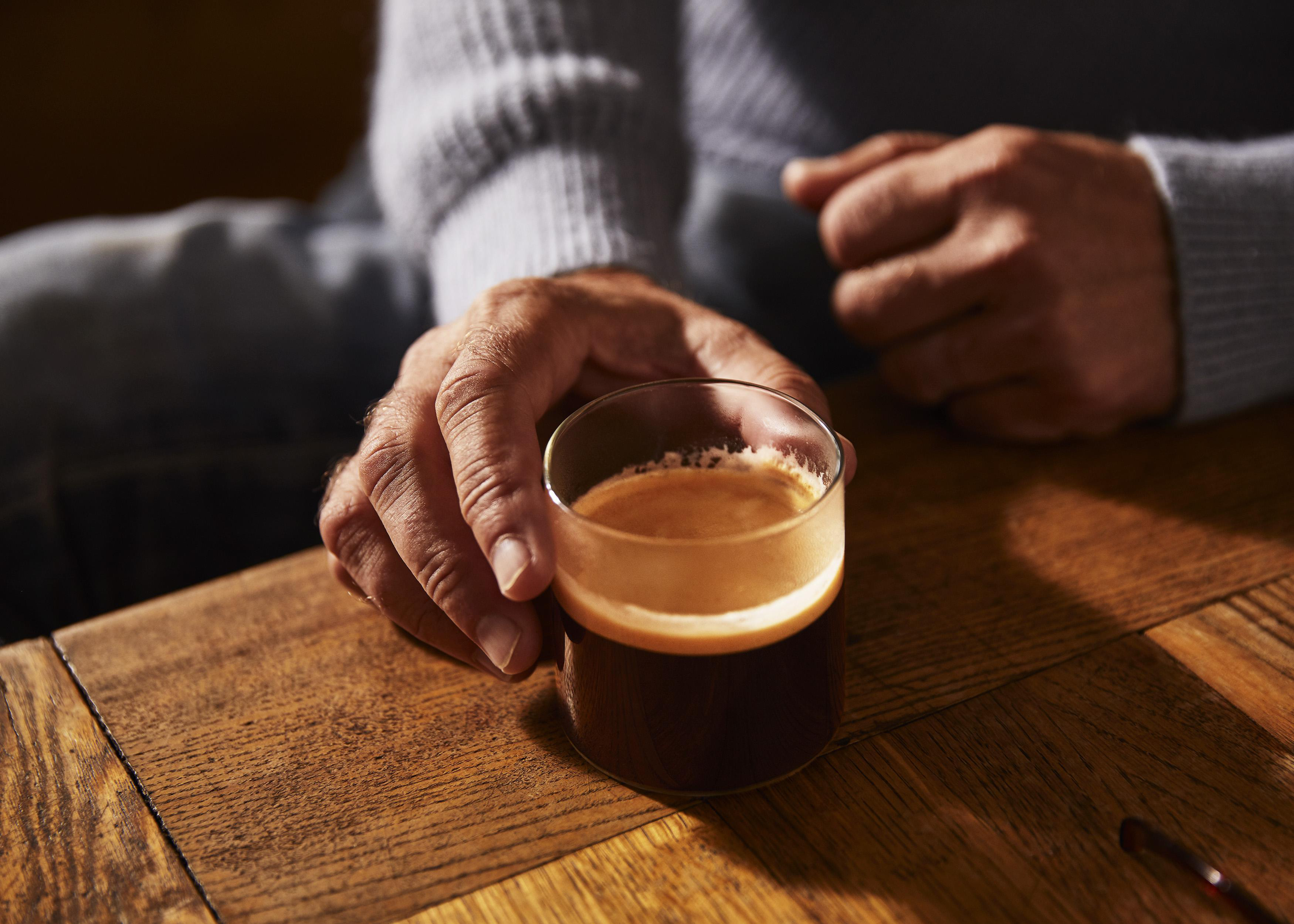 Hand, die ein Glas Espresso auf einem Holztisch hält.