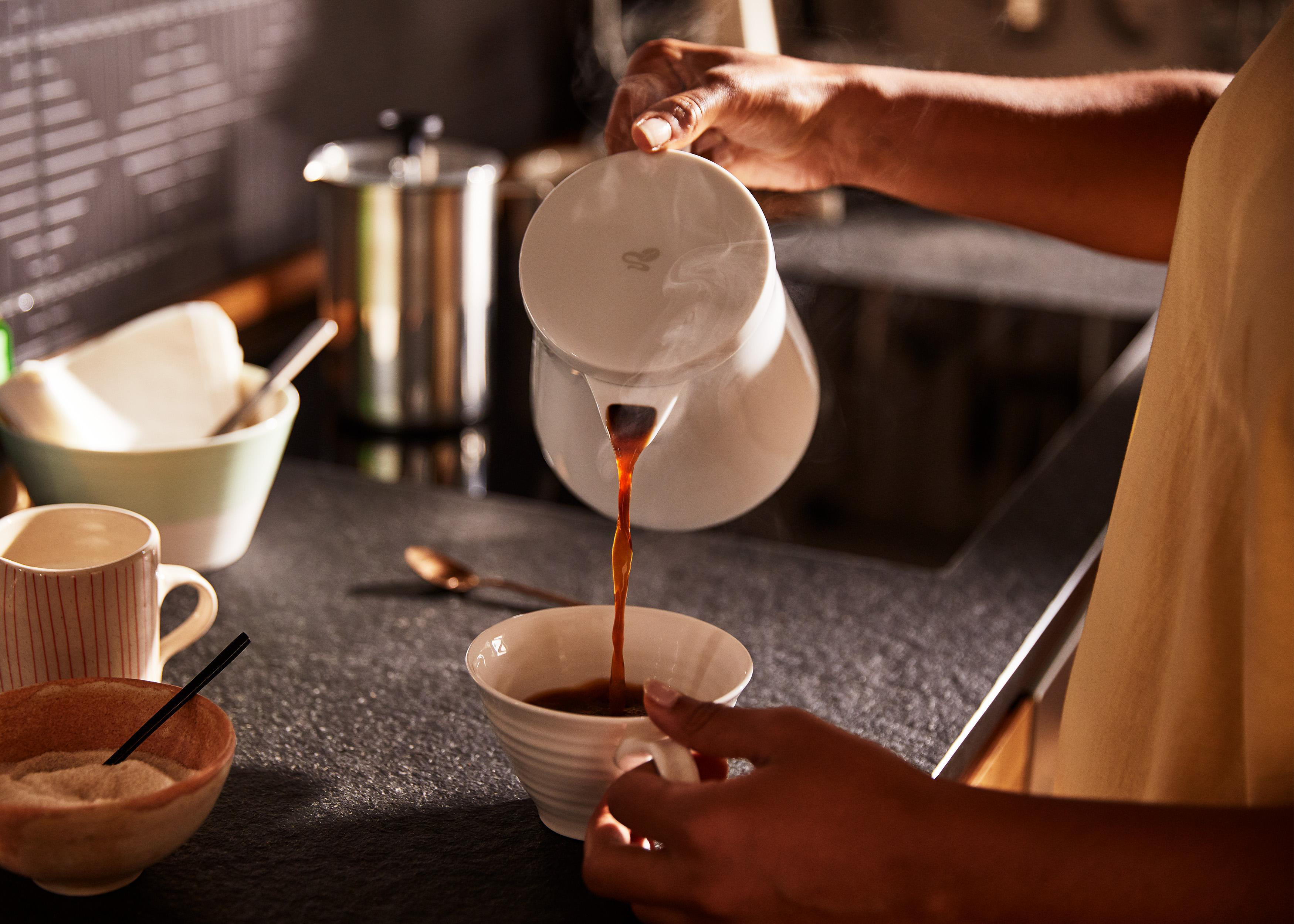 Person gießt Kaffee aus weißer Karaffe in eine Tasse, weitere Kaffeeartikel auf der Theke.