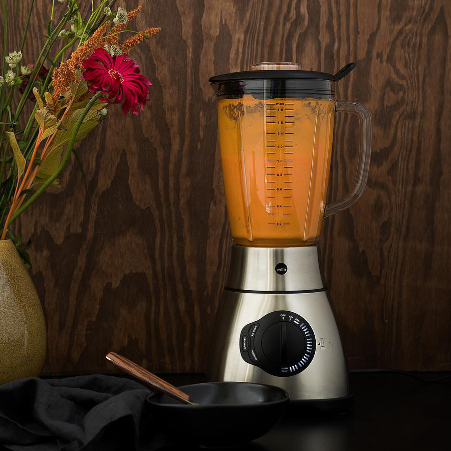 Mixer mit orangefarbener Flüssigkeit, neben einer Vase mit Blumen, einer schwarzen Schüssel und einem Holzlöffel.