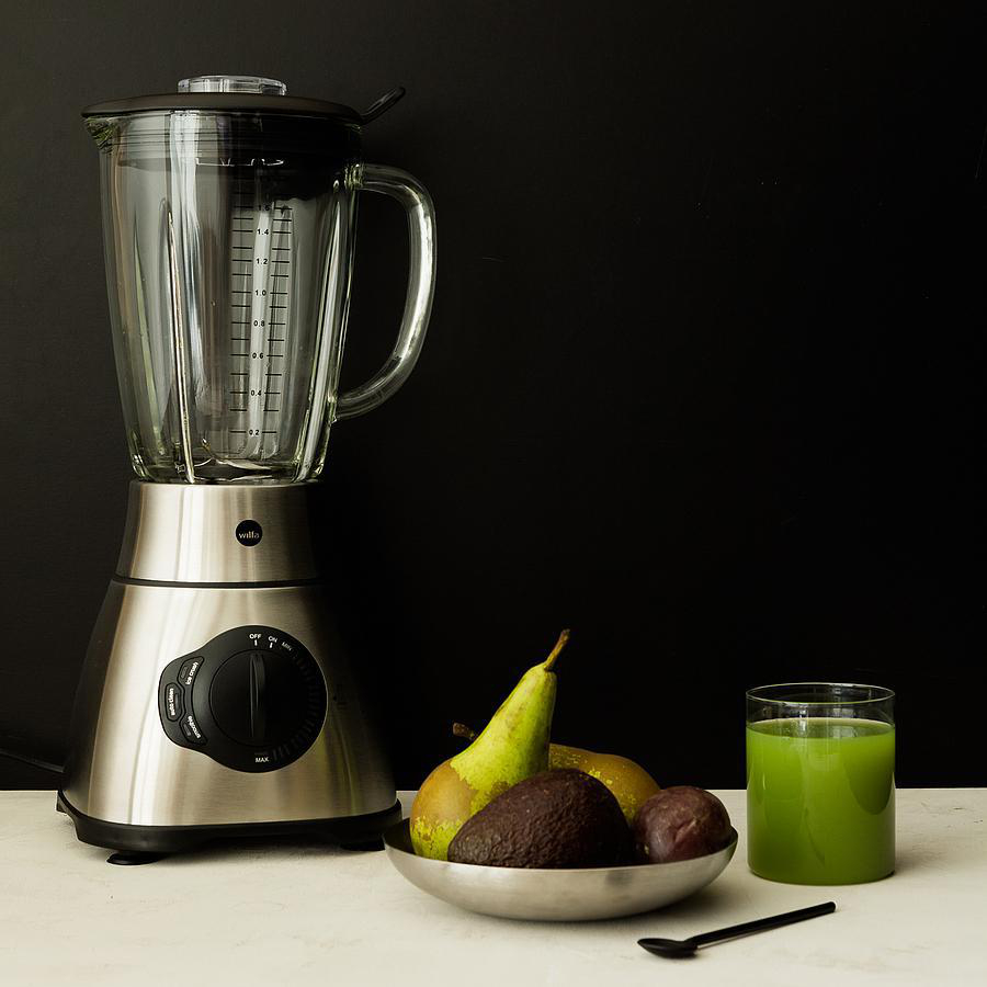 Ein Mixer mit Früchten und ein Glas grüner Saft auf weißer Oberfläche vor schwarzem Hintergrund.