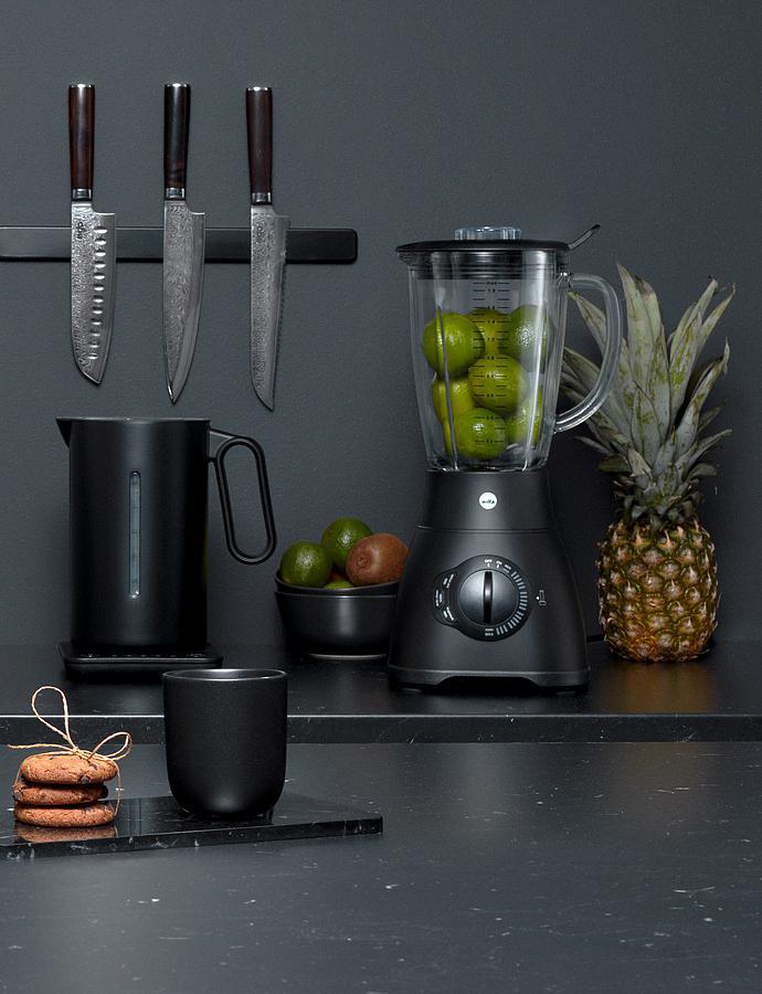 Schwarze Küchenszene mit Messern, einem Mixer mit Limetten und einer Ananas. Es gibt einen schwarzen Krug und eine Tasse.