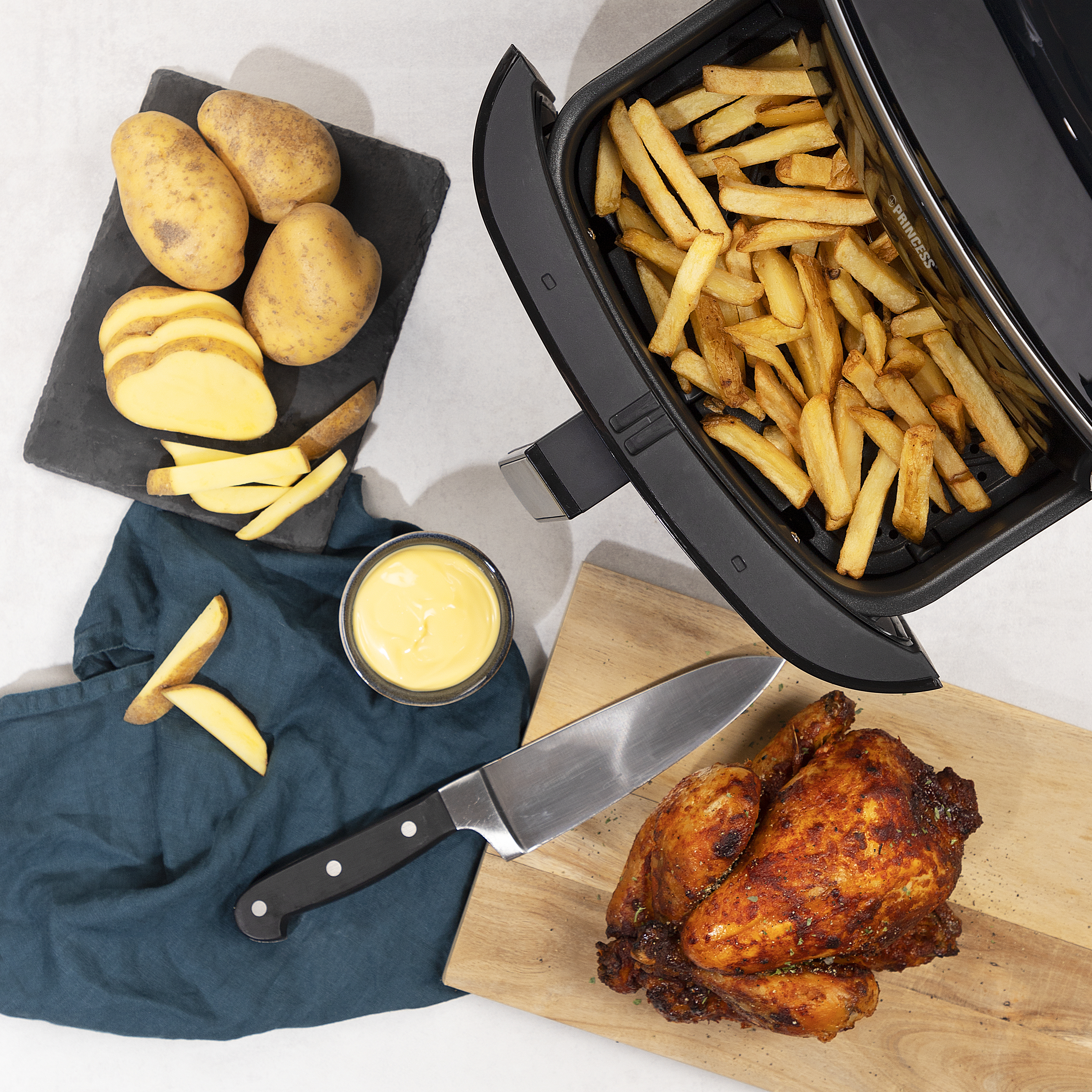 Draufsicht: Fritteuse mit Pommes, gebratenem Huhn, Kartoffeln, Dip und Messer auf weißer Fläche.