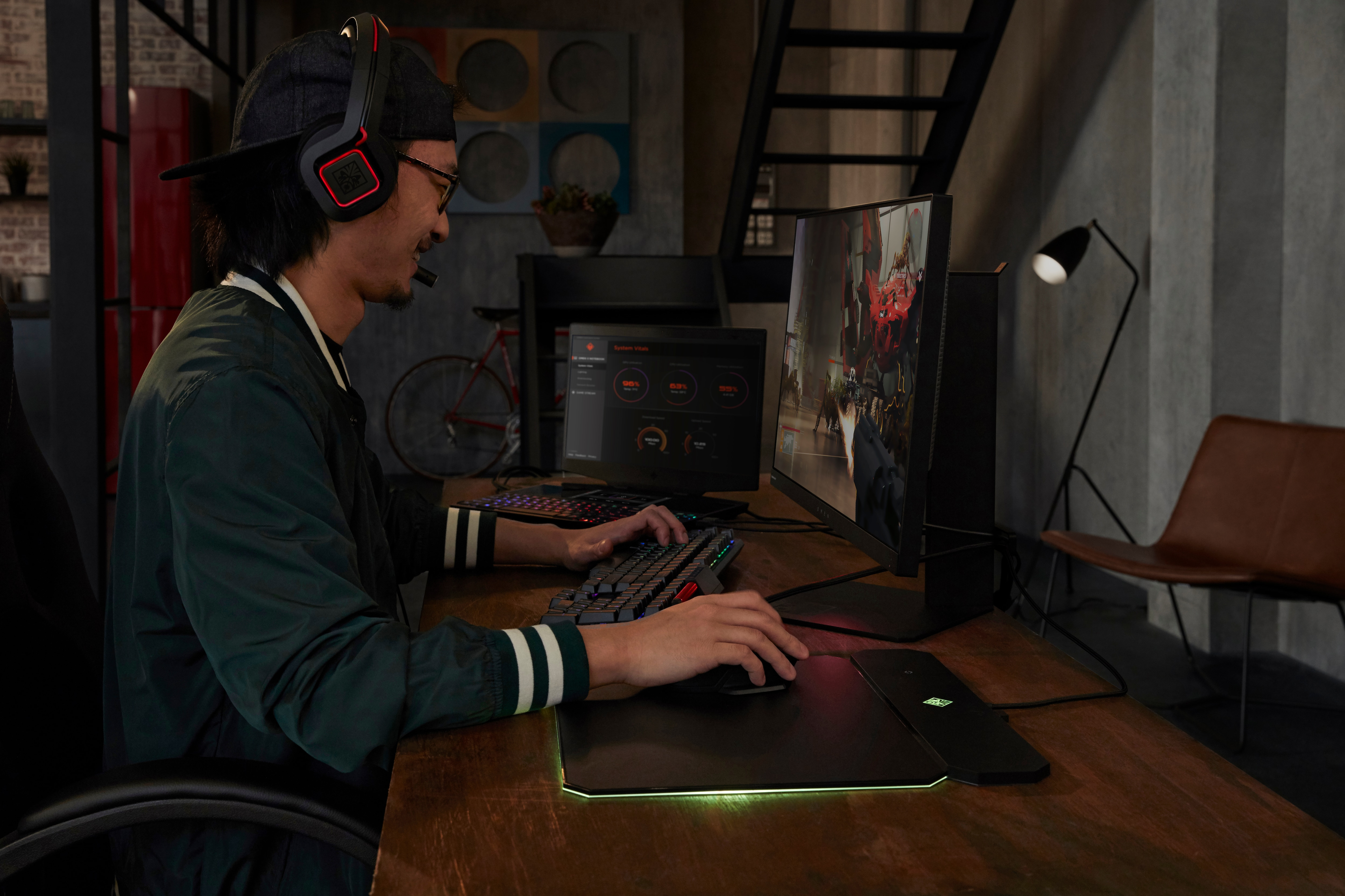 Un hombre con auriculares y gorra, jugando en un escritorio con dos monitores, teclado y ratón, habitación oscura.