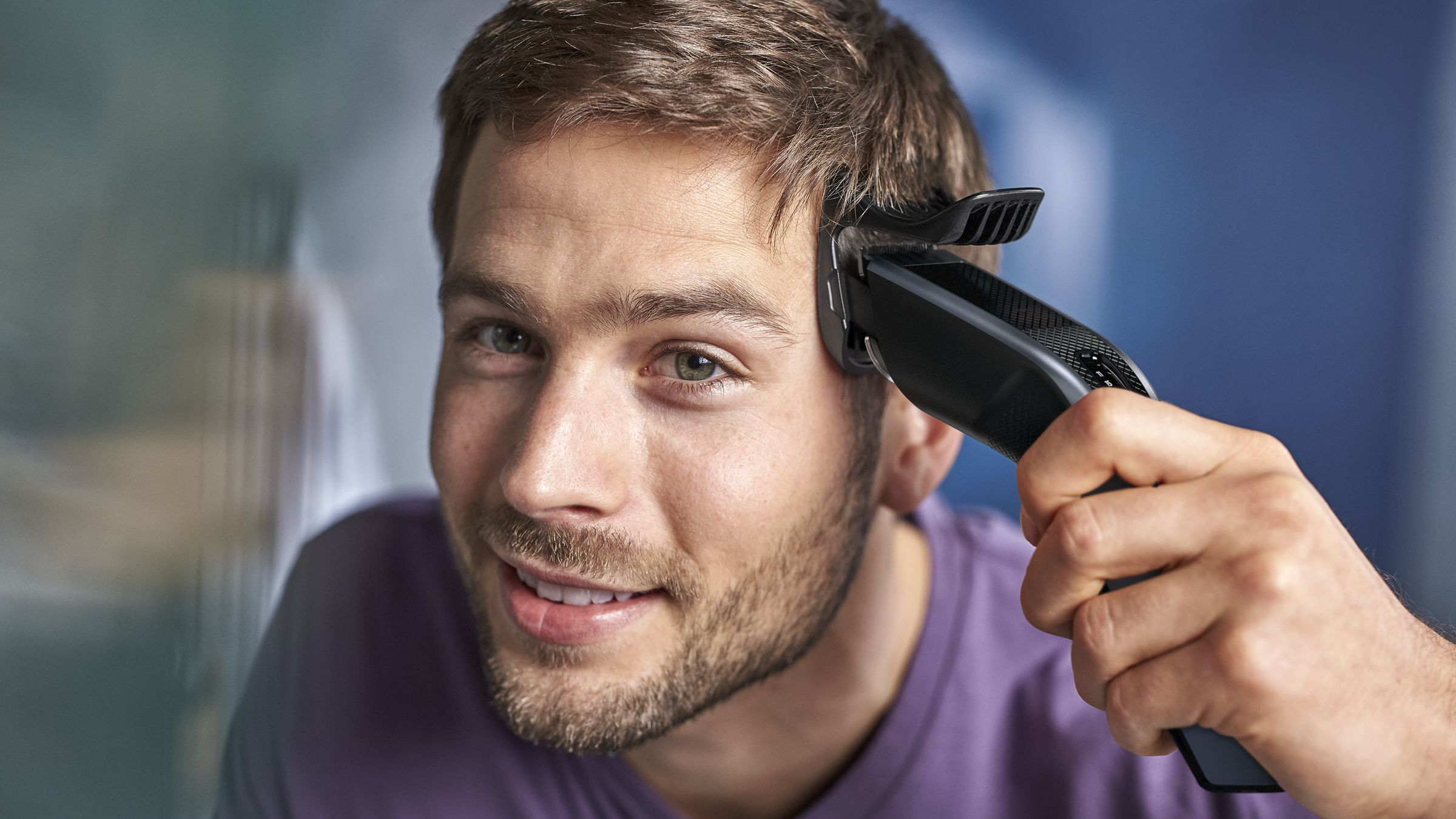 Hombre usando una cortadora de pelo eléctrica negra. Sonriendo, fondo claro.