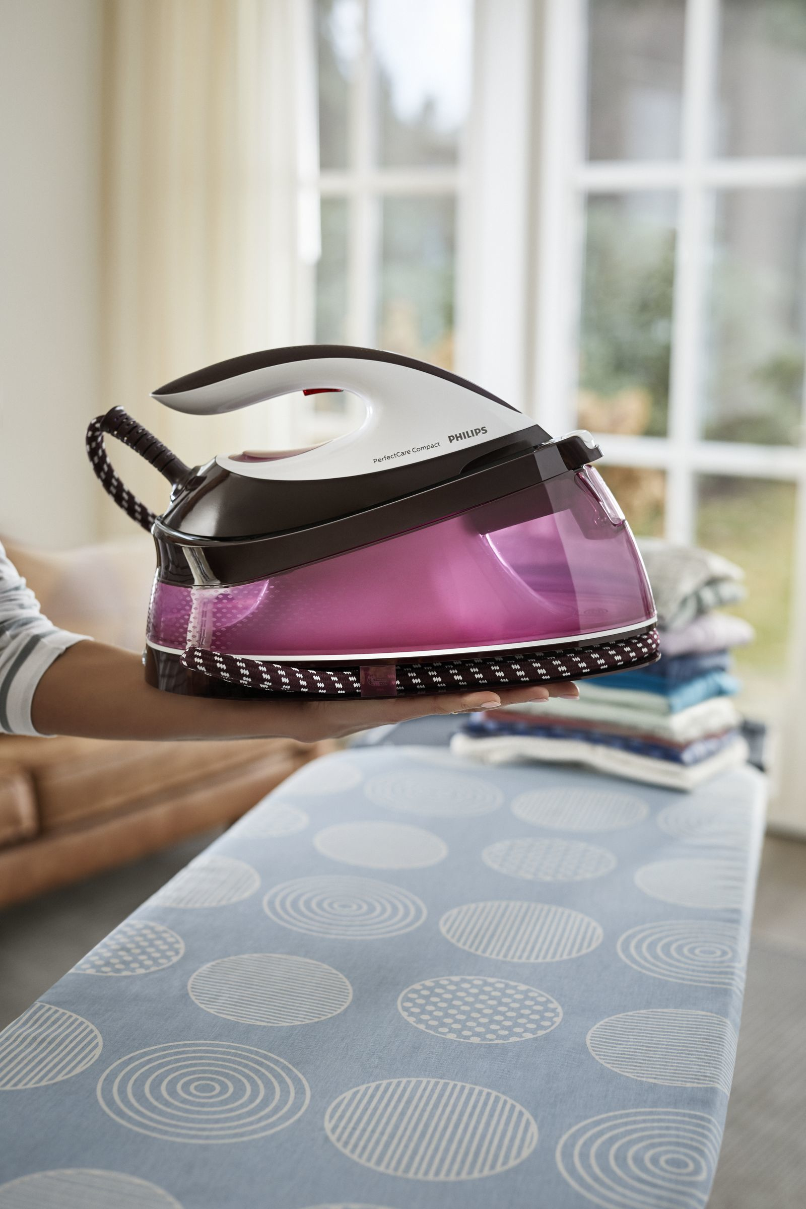 Mujer sosteniendo plancha Philips rosa y blanca cerca de tabla de planchar con ropa.