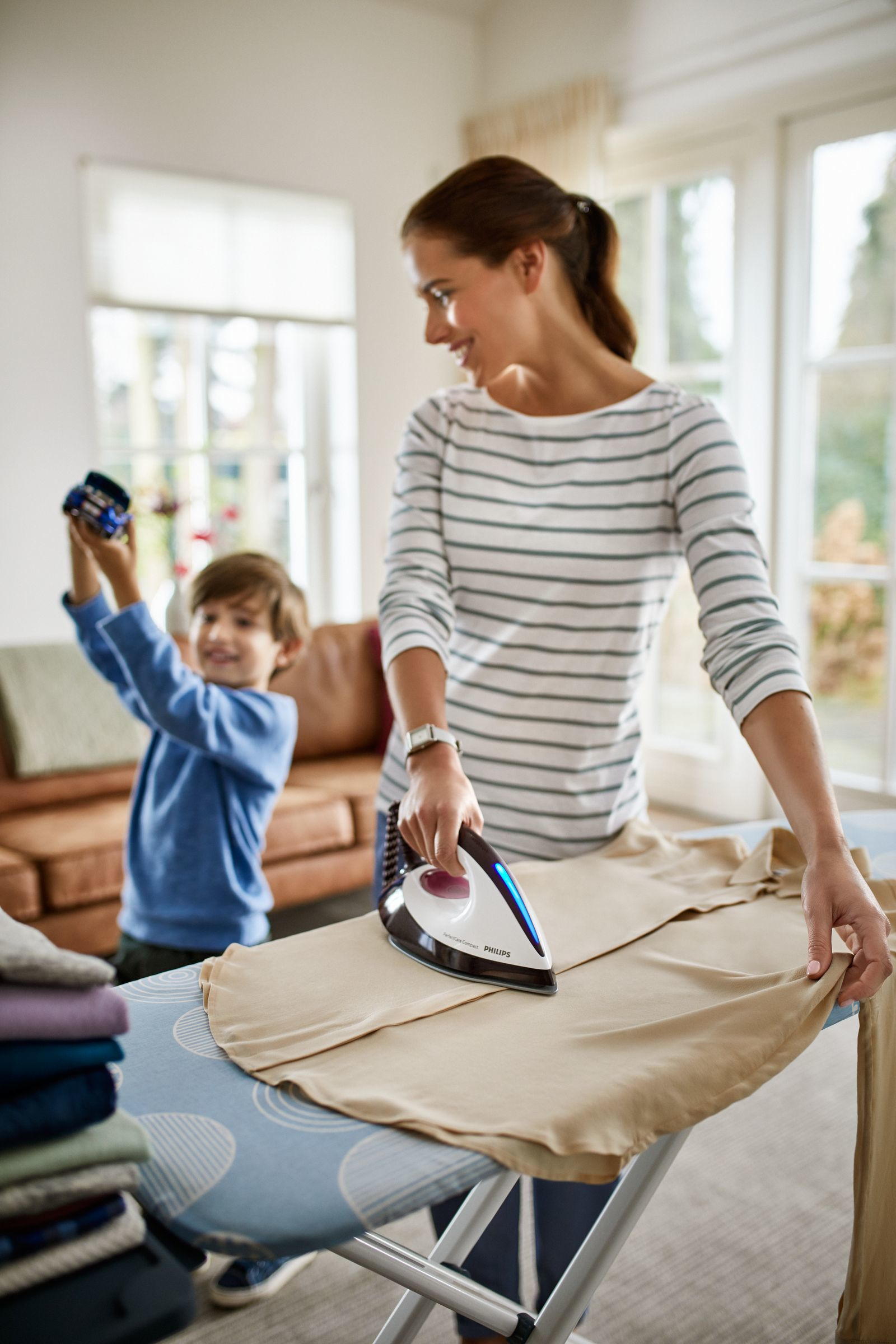 Una mujer planchando ropa en una tabla de planchar, con un niño sosteniendo ropa en el fondo.