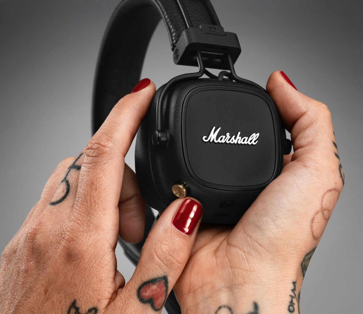 Hands with tattoos hold a black Marshall headphone, red nail polish, and gold button.