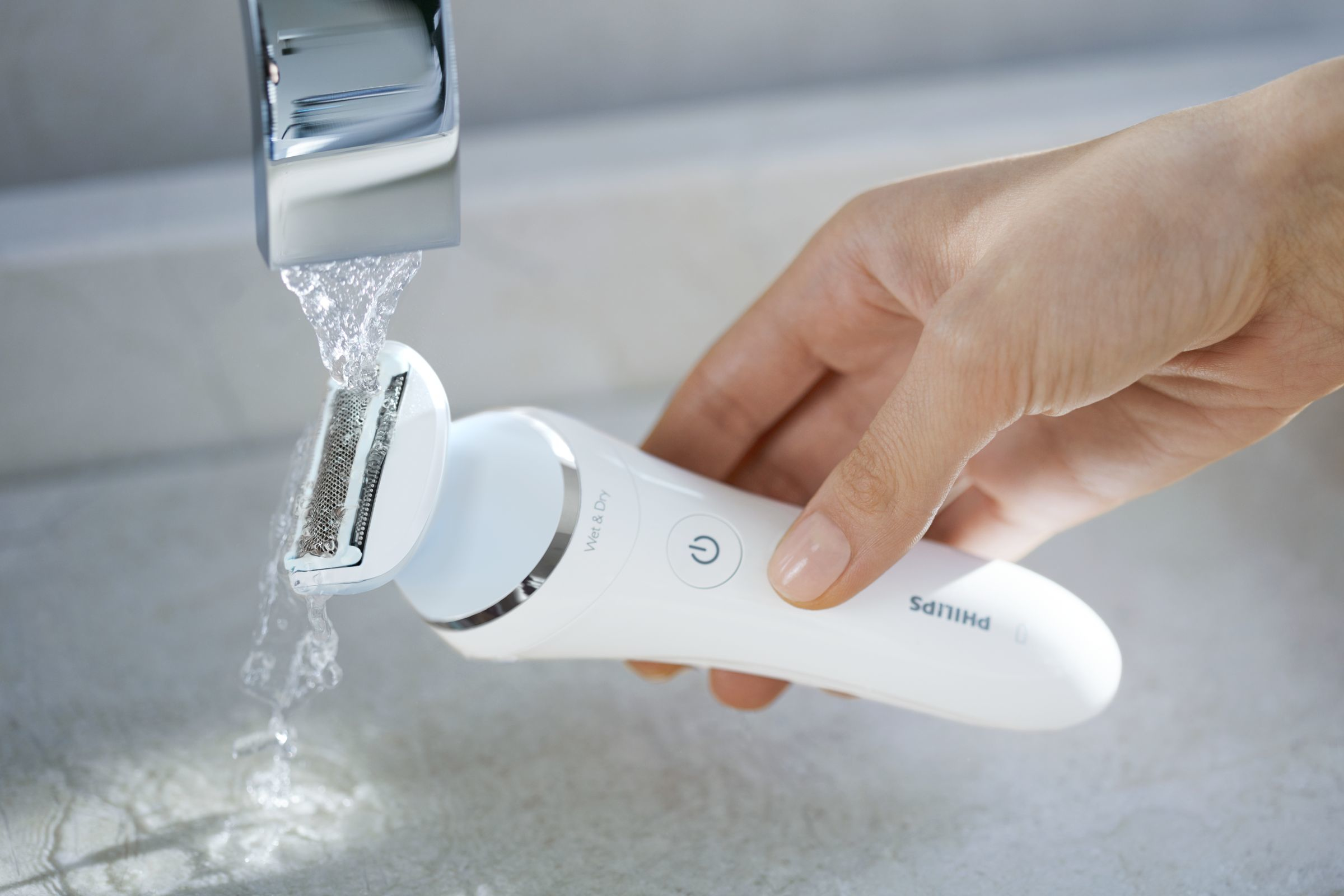 La mano de una mujer enjuagando una afeitadora Philips húmeda y seca bajo agua corriente.
