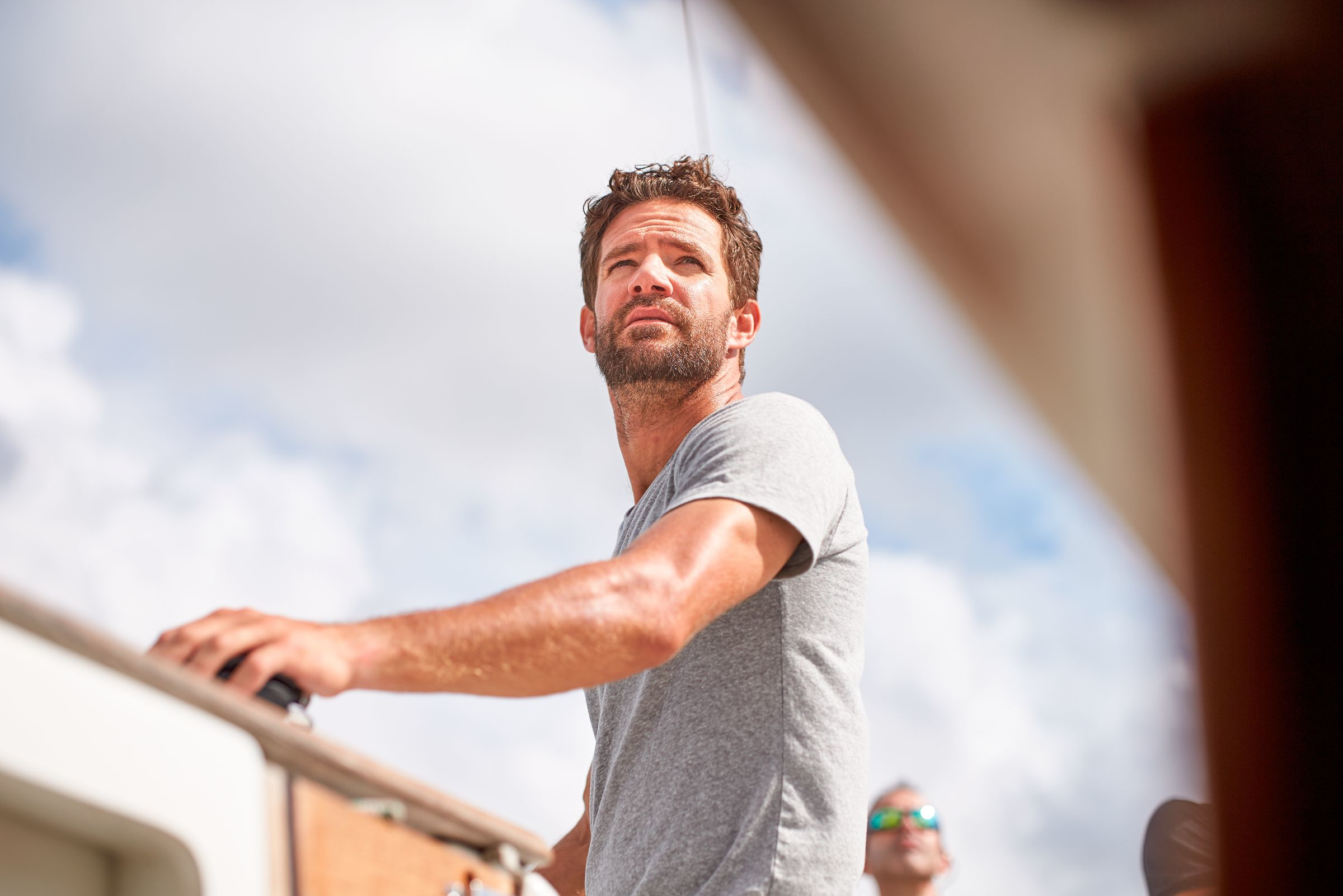 Un hombre mirando hacia arriba, en un barco. Lleva una camisa gris, con un cielo brillante en el fondo.
