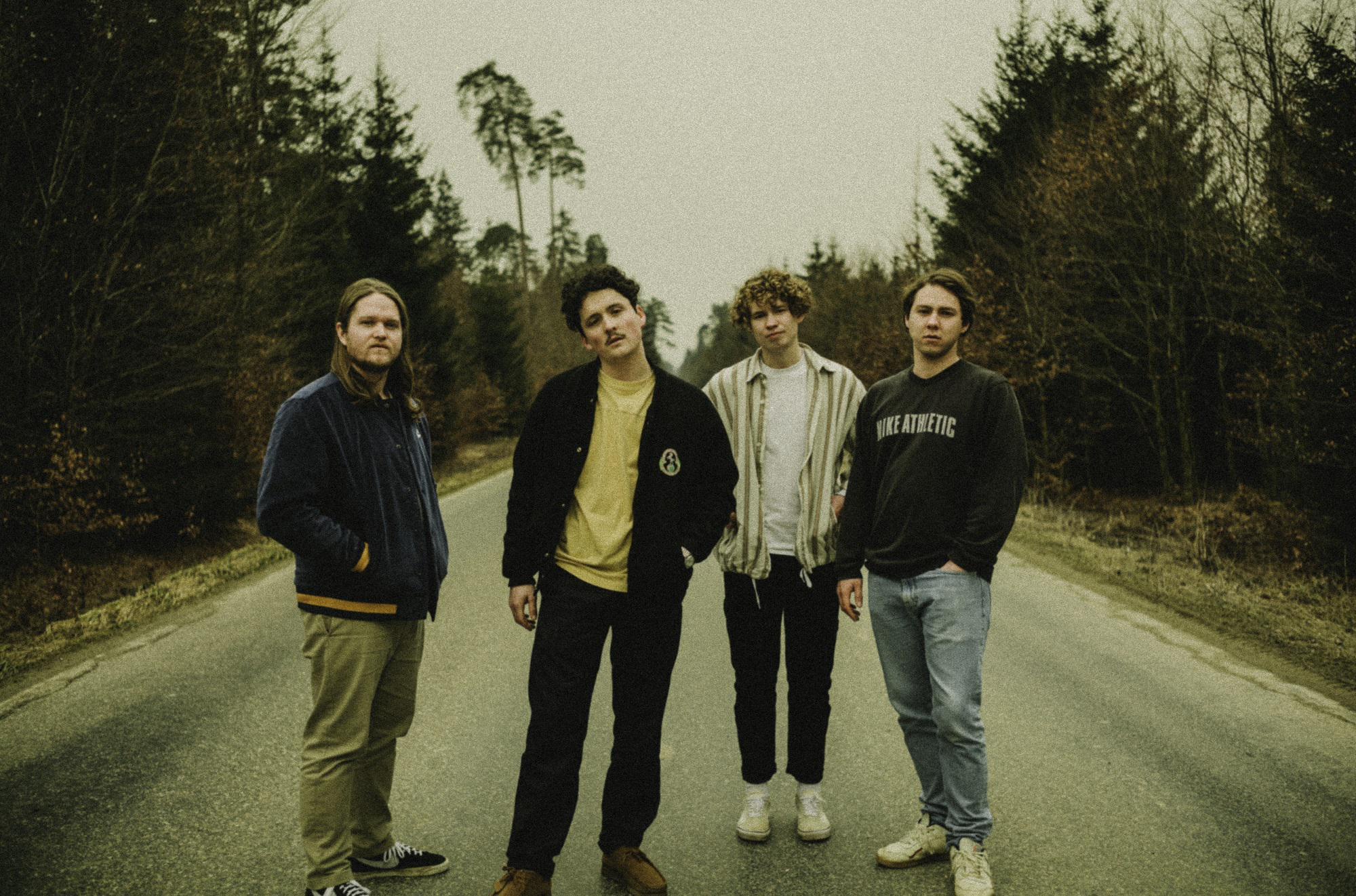 Eine Band aus vier Personen steht auf einer Straße in einem Wald.