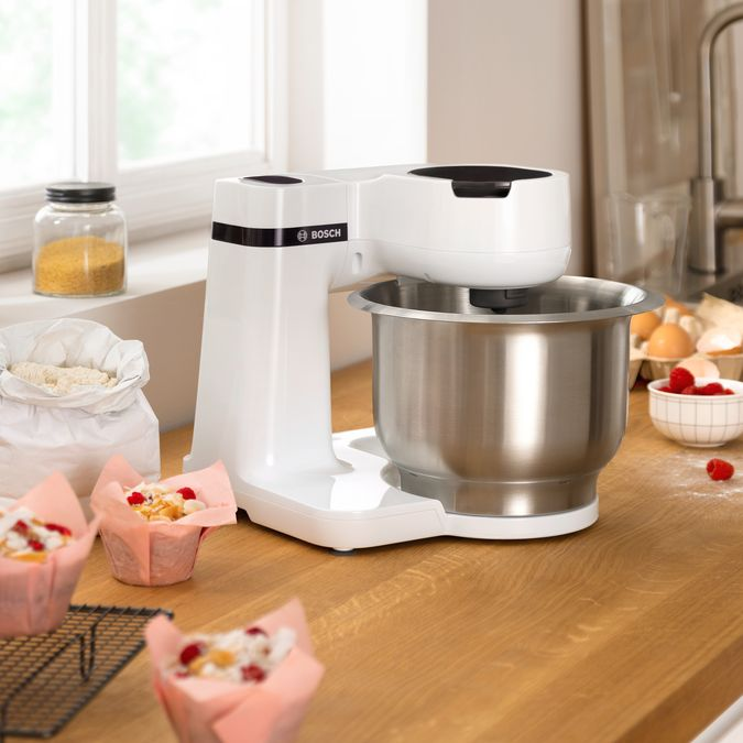 Fehér robotgép rozsdamentes acél tálal, muffinokkal és hozzávalókkal a pulton.
