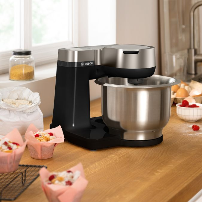 Egy fekete-ezüst Bosch konyhai robotgép rozsdamentes acél tálkával, muffinokkal és hozzávalókkal.
