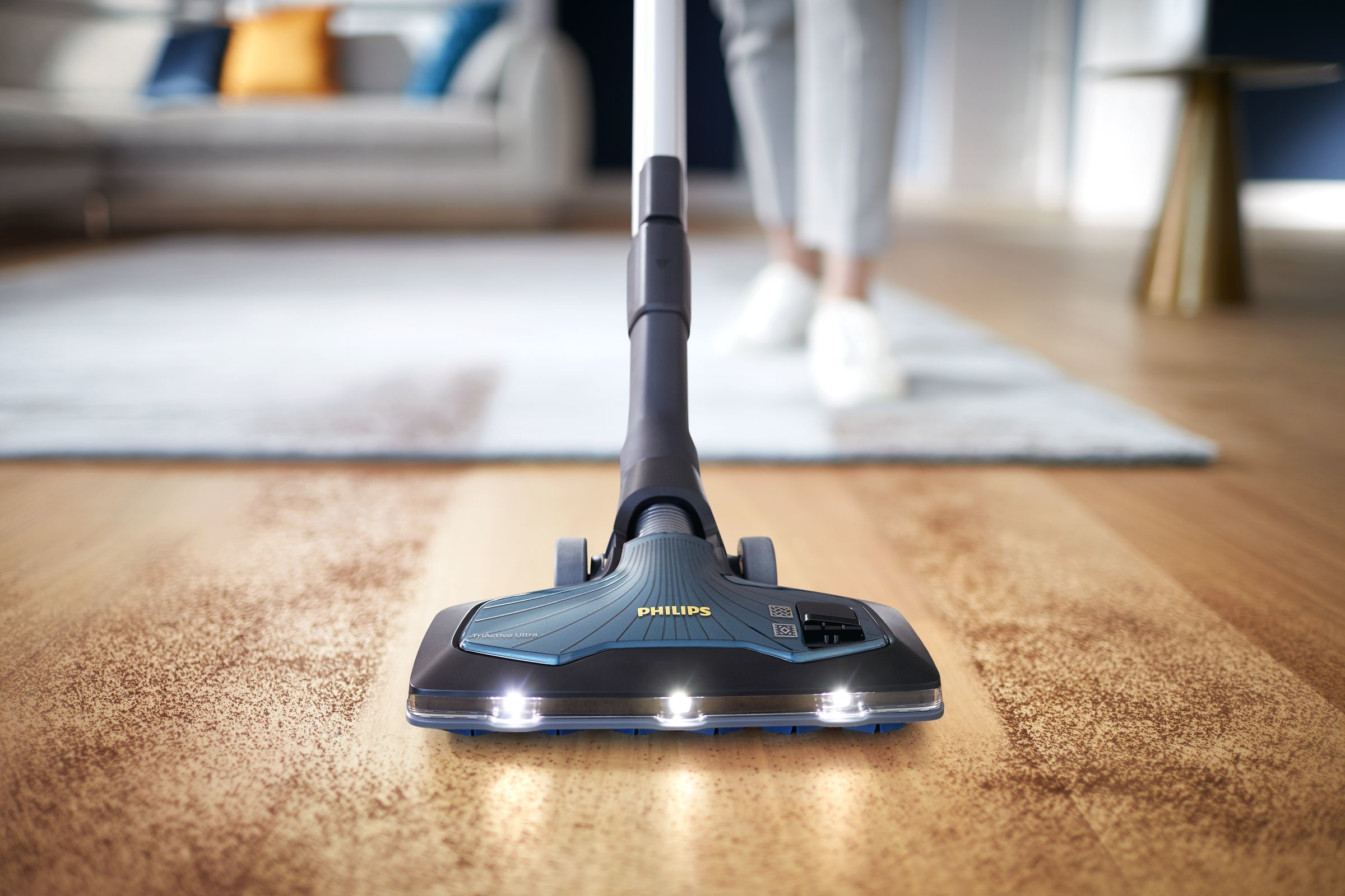 Ahşap zeminde elektrikli süpürge başlığının yakın çekimi, arka planda kişi, ışıklar açık.