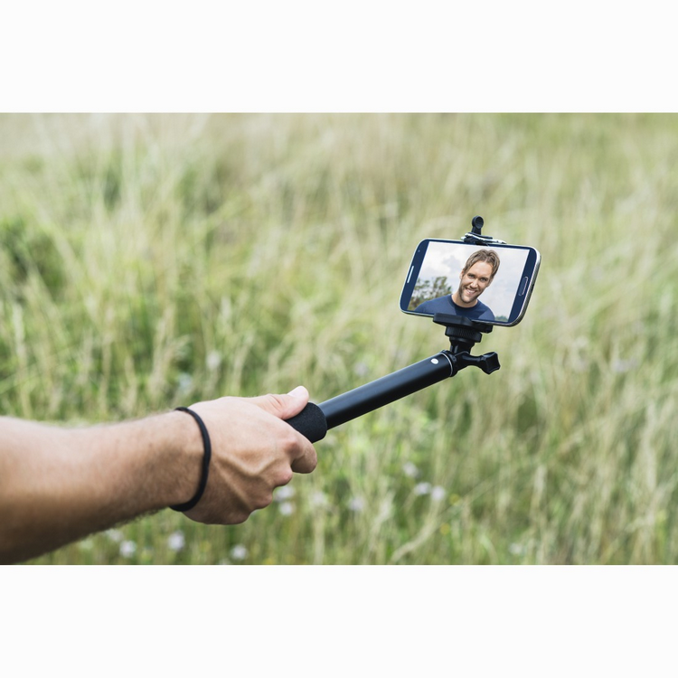 Hombre sosteniendo un palo selfie con un teléfono que muestra su cara en un campo verde.