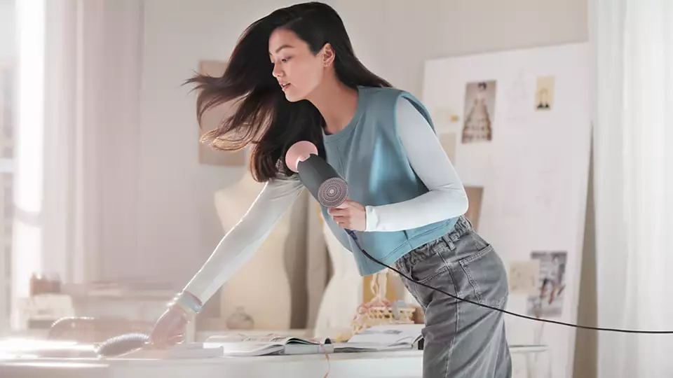 Une femme se coiffe avec un sèche-cheveux, dans une pièce avec un bureau.