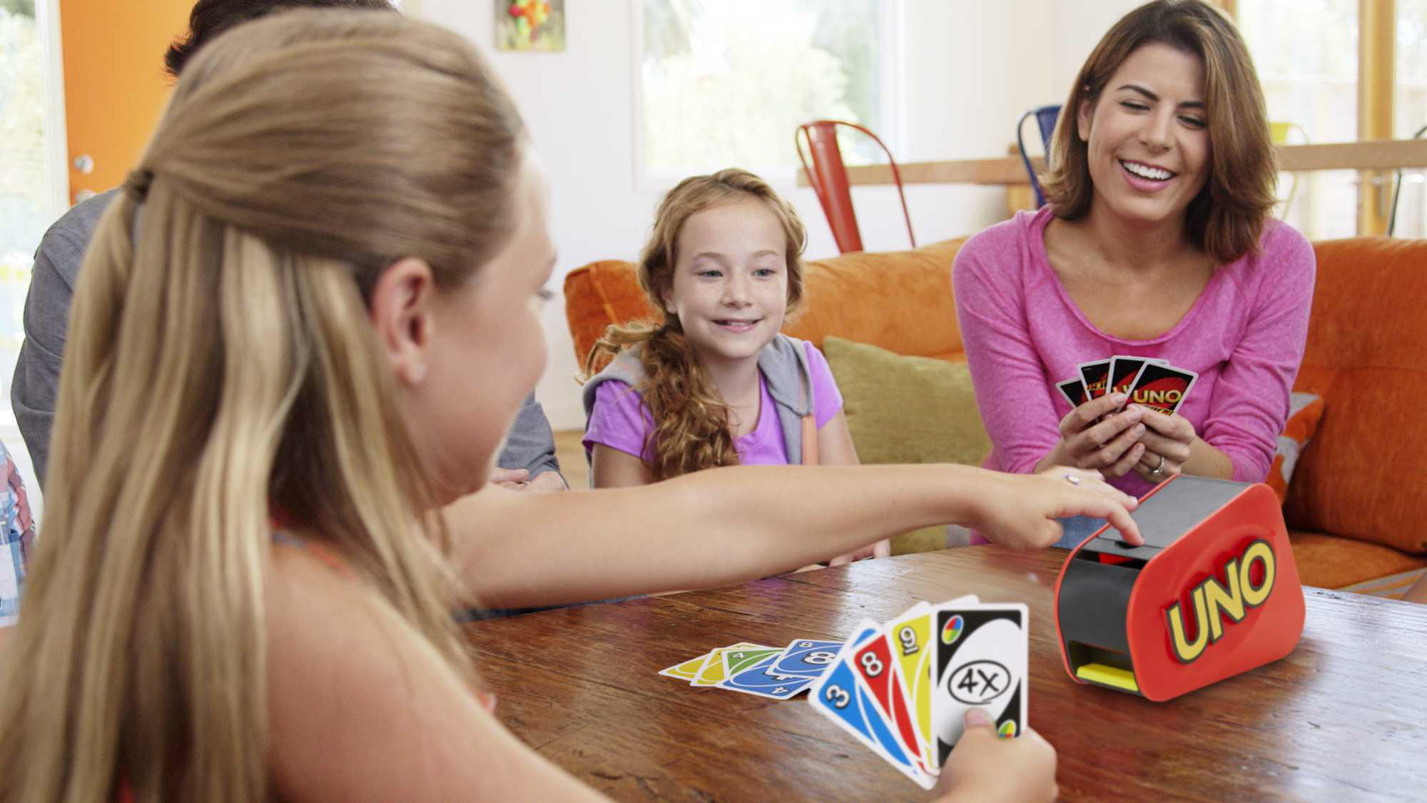 Familie spielt UNO. Ein Mädchen spielt Karten. Eine rote UNO-Box steht auf dem Tisch.