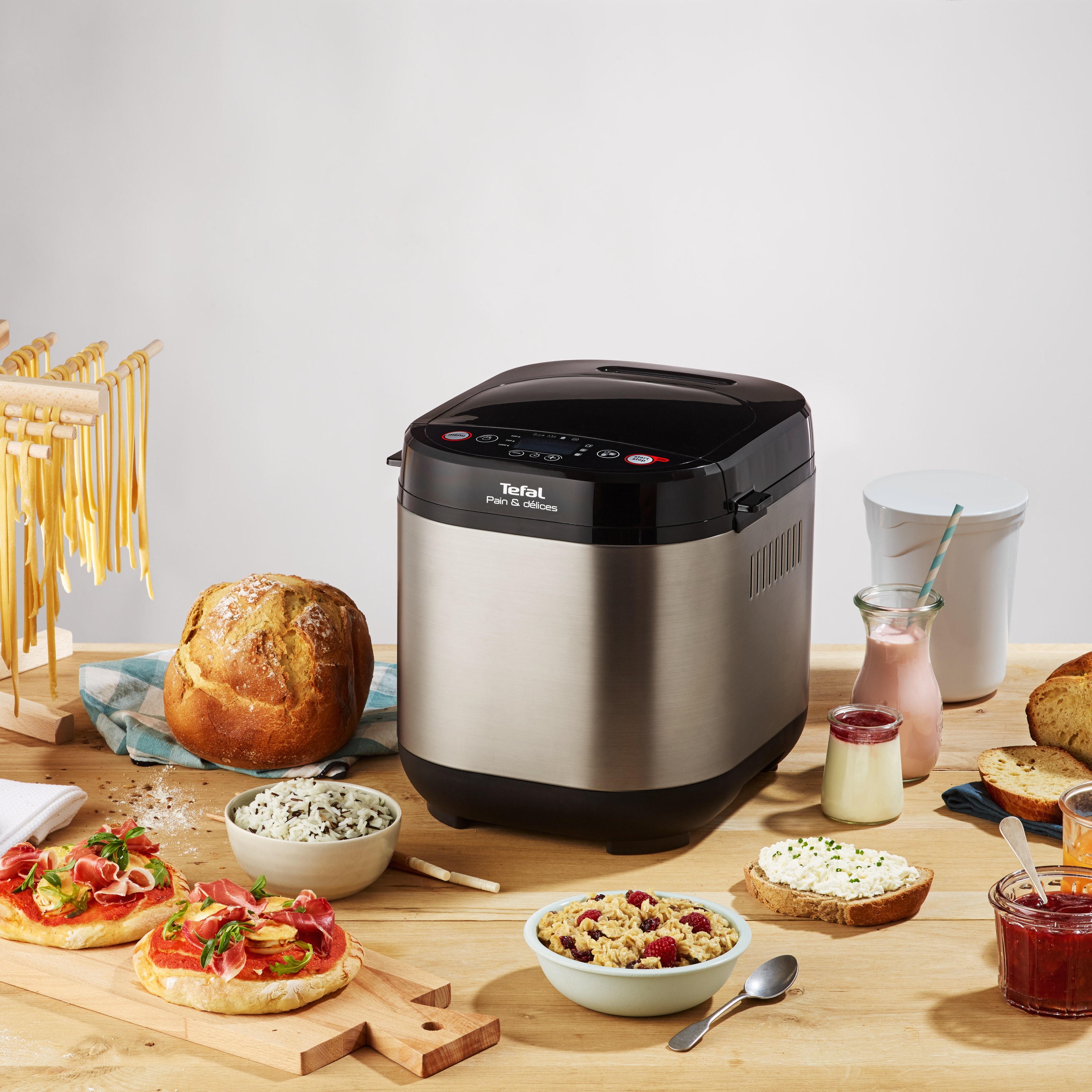 Eine Brotbackmaschine steht auf einem Holztisch, zusammen mit anderen Lebensmitteln wie Brot und Tomaten.