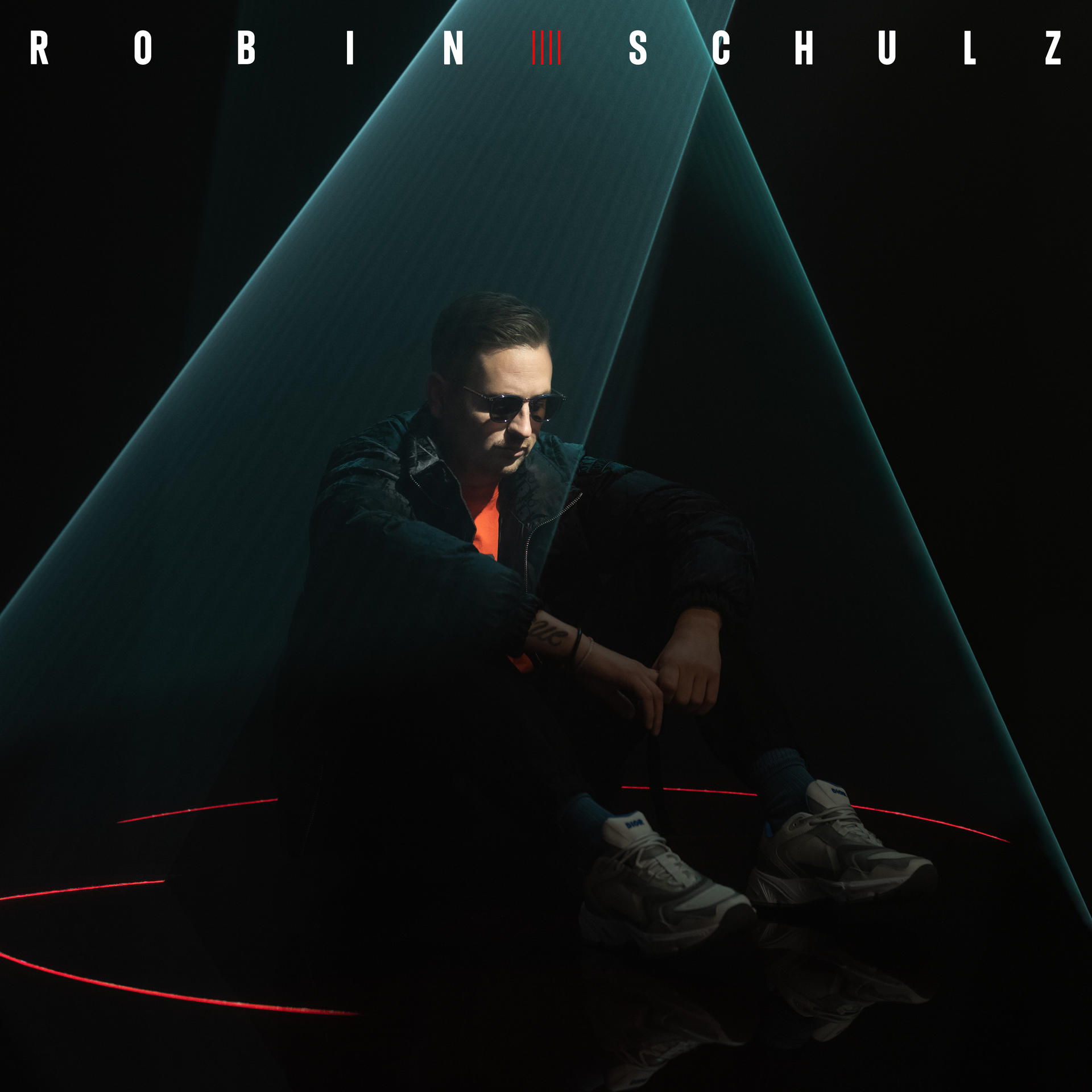 Robin Schulz assis, portant des lunettes de soleil, devant un fond sombre avec des lumières néon bleues et rouges.