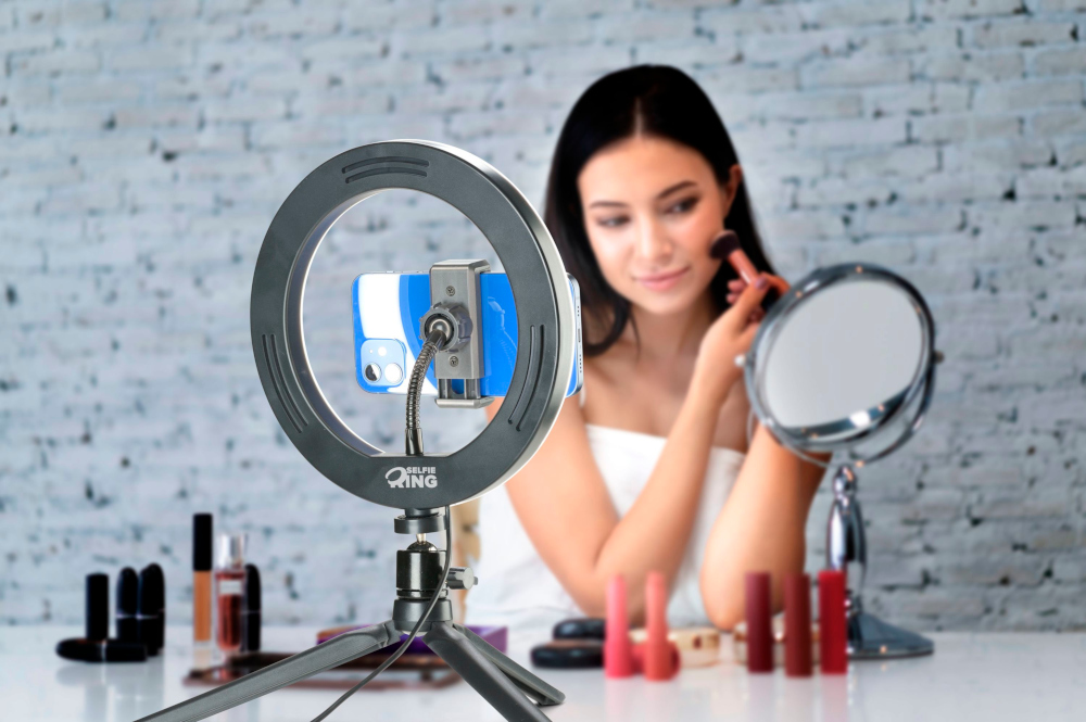 Een vrouw brengt make-up aan. Een selfie ringlicht en een spiegel staan voor. Lippenstift en andere items staan op de tafel.