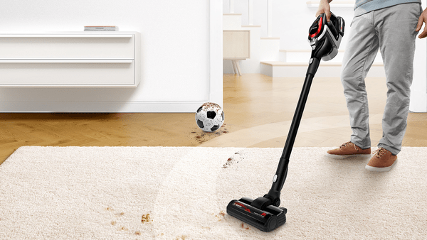 Ein Mann benutzt einen kabellosen Staubsauger in Schwarz und Rot auf einem Teppich. Ein schmutziger Fußball liegt am Boden.