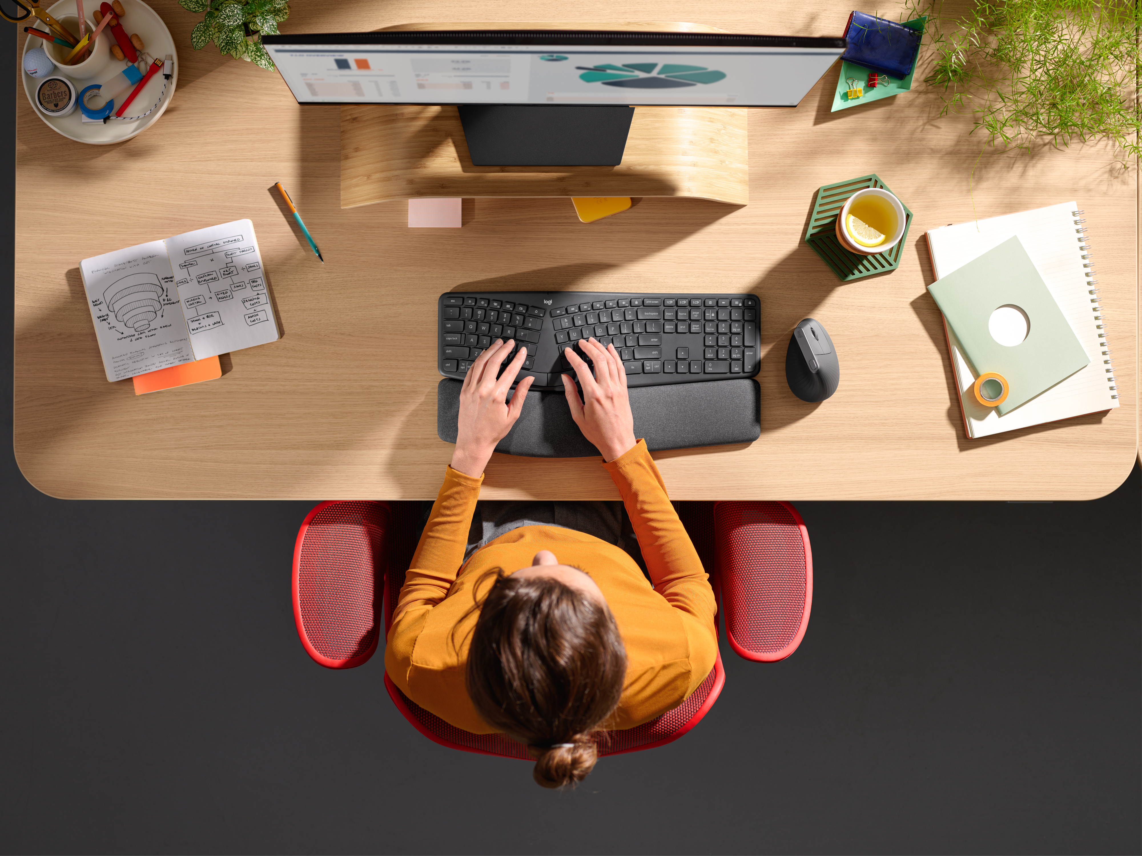 Una donna a una scrivania con tastiera, monitor, mouse e forniture per ufficio. Indossa un top arancione.