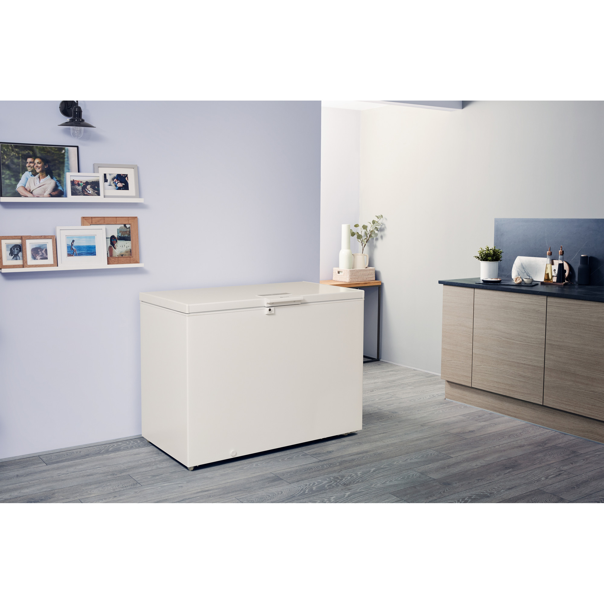 Eine weiße Truhen-Gefriertruhe in einem Raum. Bilder an der Wand. Holzschrank rechts.
